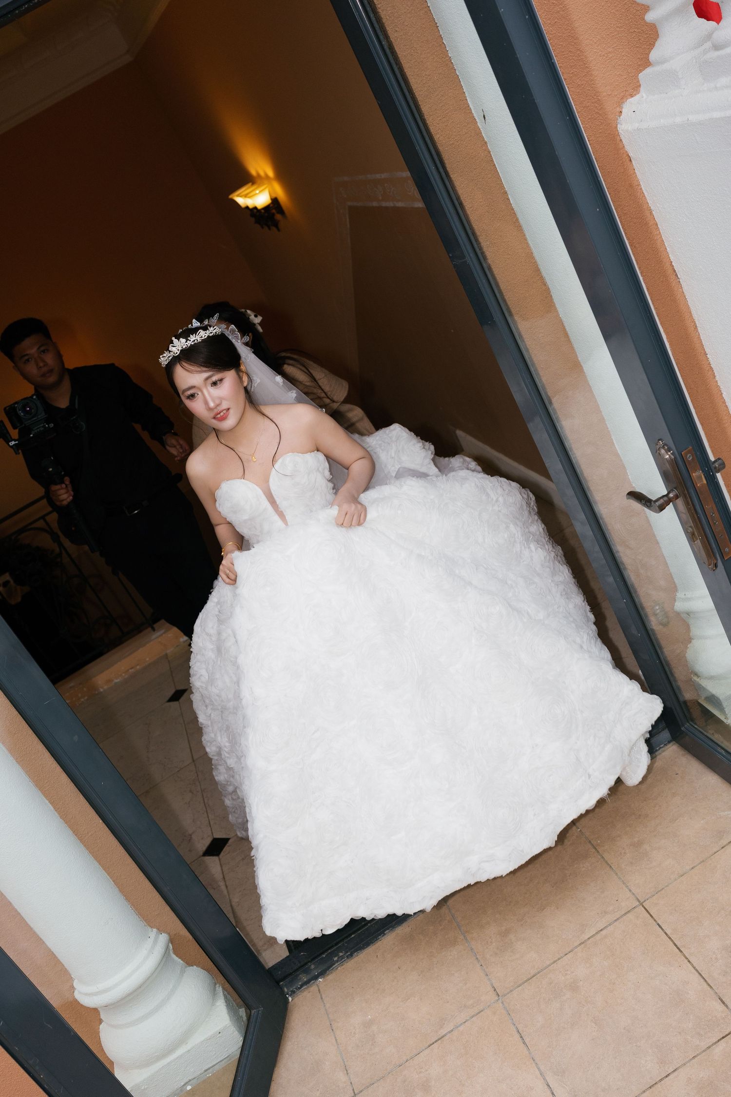 A bride in a white ball gown and tiara walks through a doorway, lifting her dress as she enters.