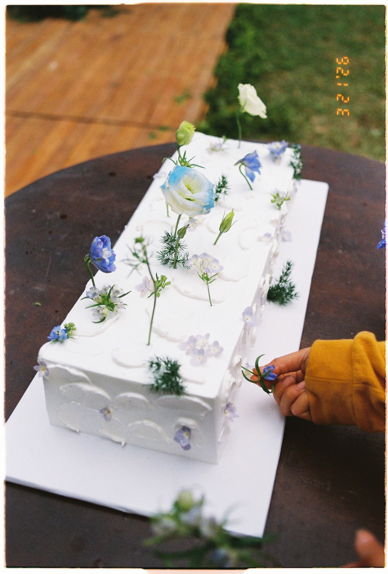 Rectangular white wedding cake decorated with blue and white flowers on a wooden table outdoors.