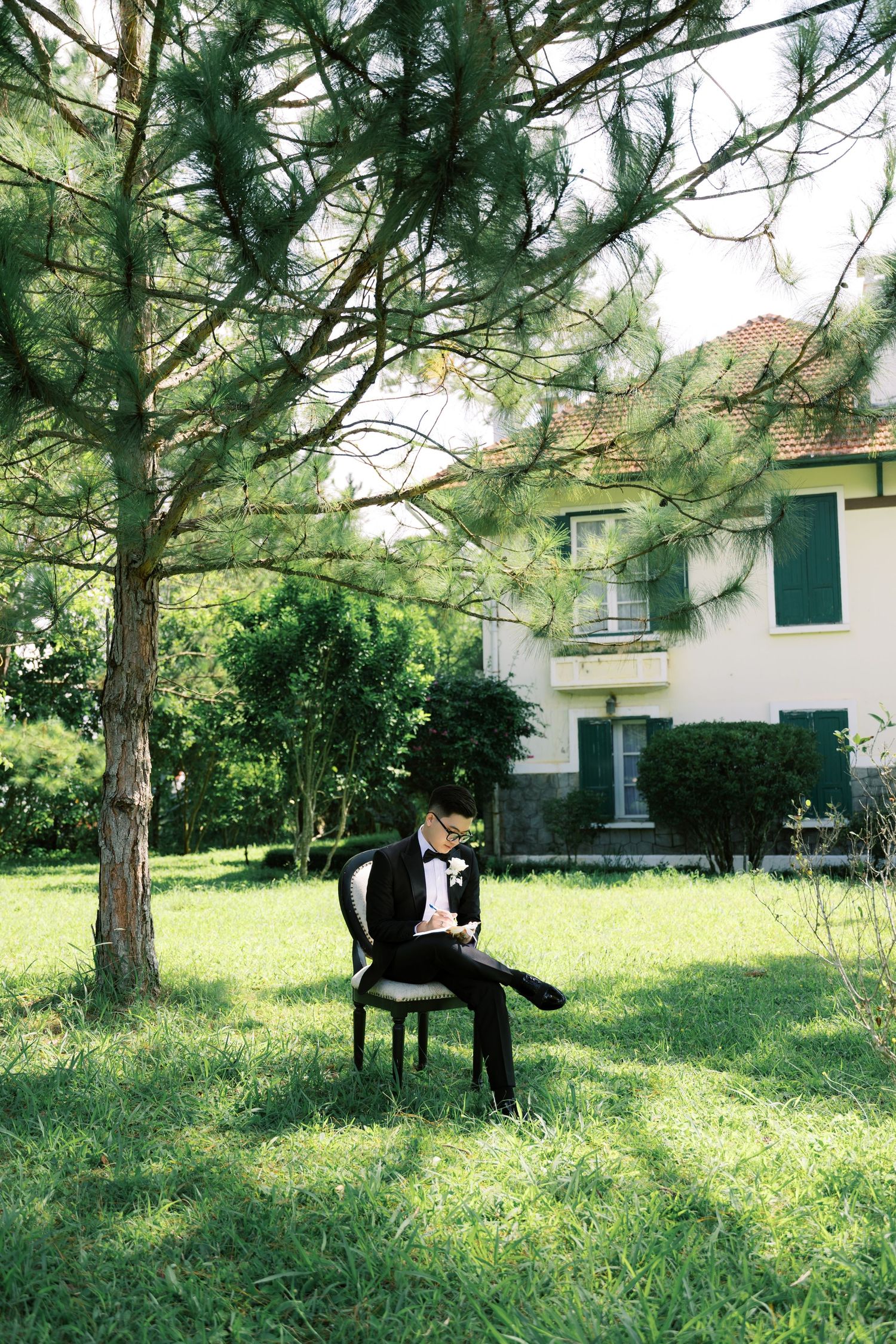 Groom in black tuxedo sitting on chair under pine tree in lush garden with white villa in background.