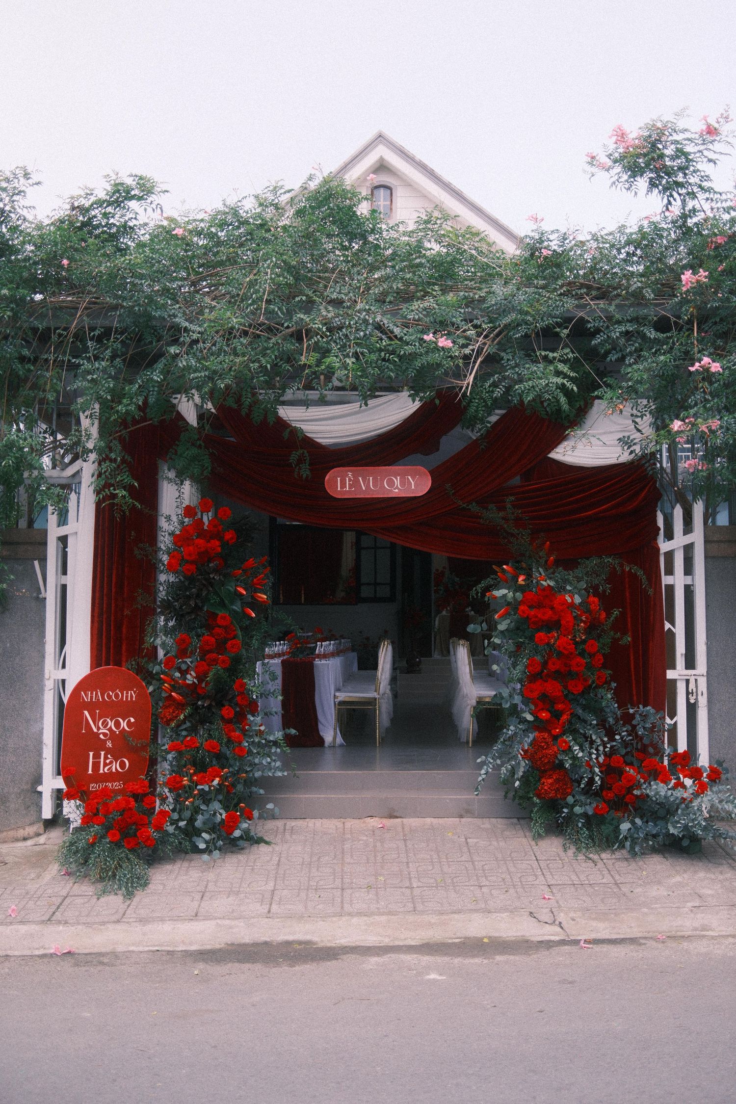 Elegant Vietnamese wedding venue entrance adorned with red roses, draped fabric, and lush greenery framing white doors.