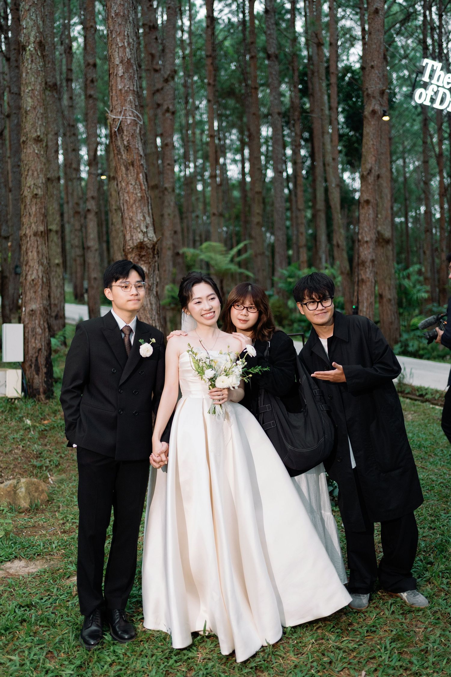 Bride in ivory ball gown holding white bouquet poses with three guests in a pine forest wedding venue.