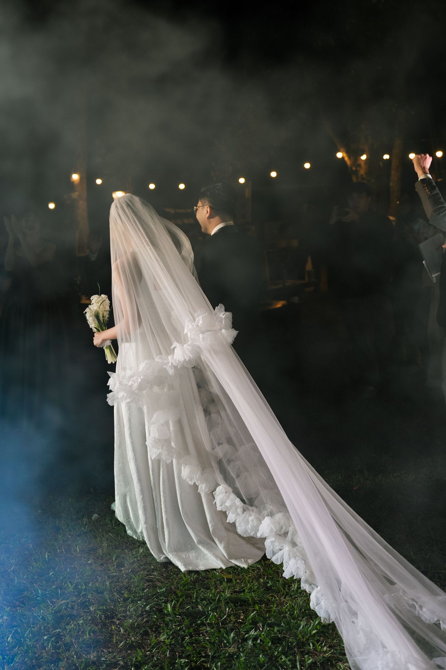 Bride in lace-trimmed veil and gown walks through dramatic smoke at outdoor nighttime wedding.
