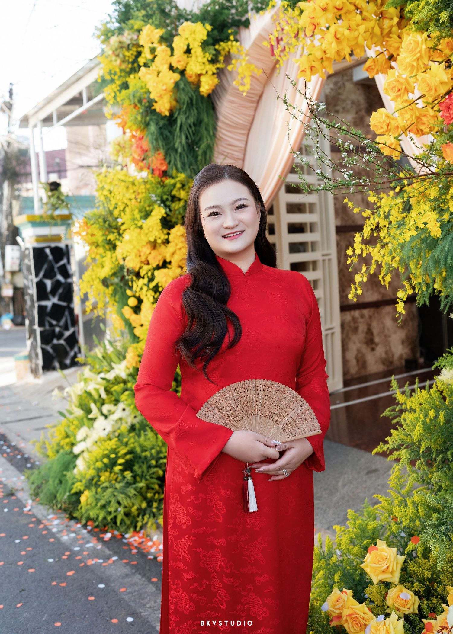 A smiling woman in a red áo dài holds a wooden fan, posing among vibrant yellow flowers outdoors.