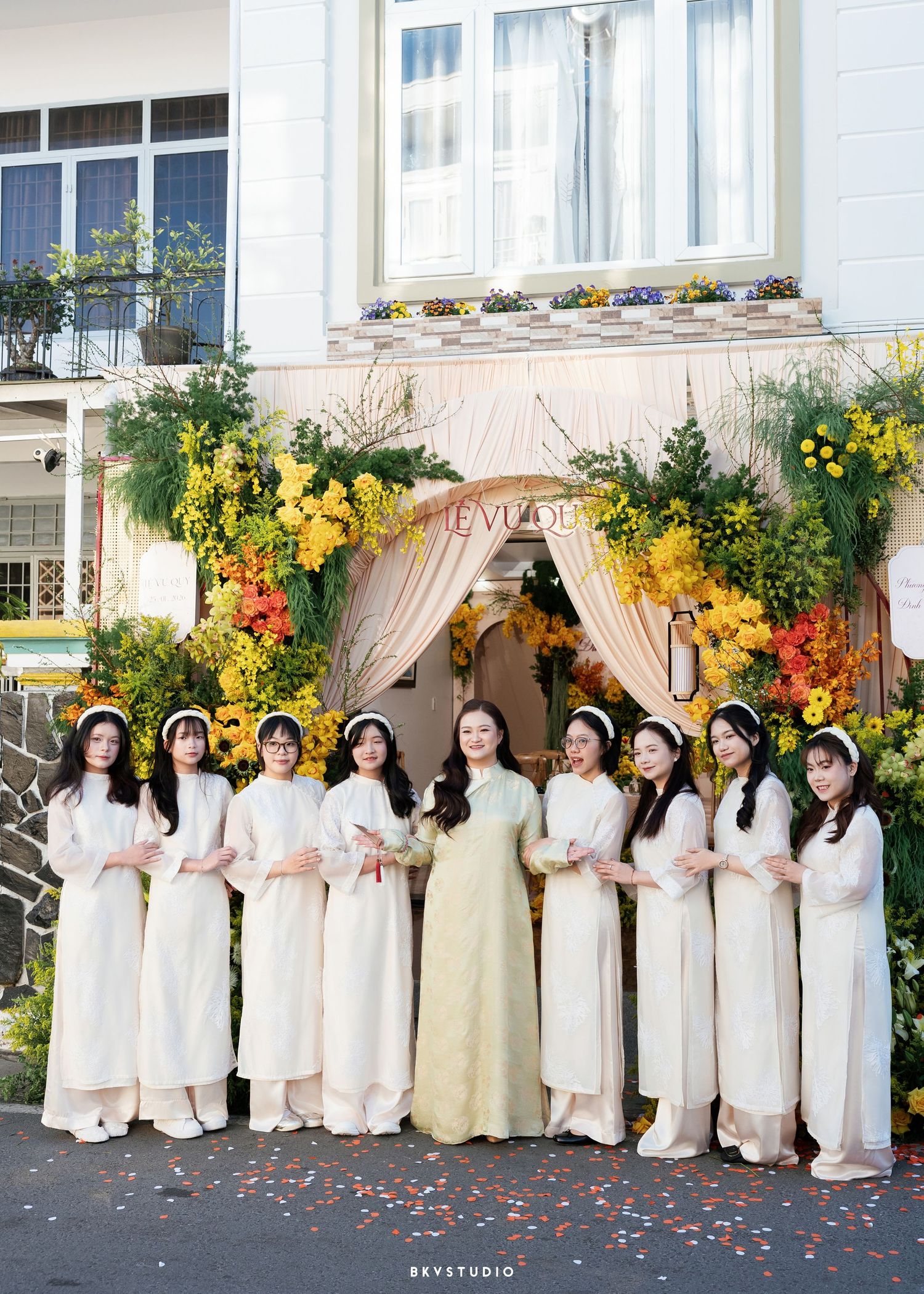 Vietnamese bridesmaids in white ao dai gowns pose with bride before colorful floral wedding arch decorated with yellow blooms.