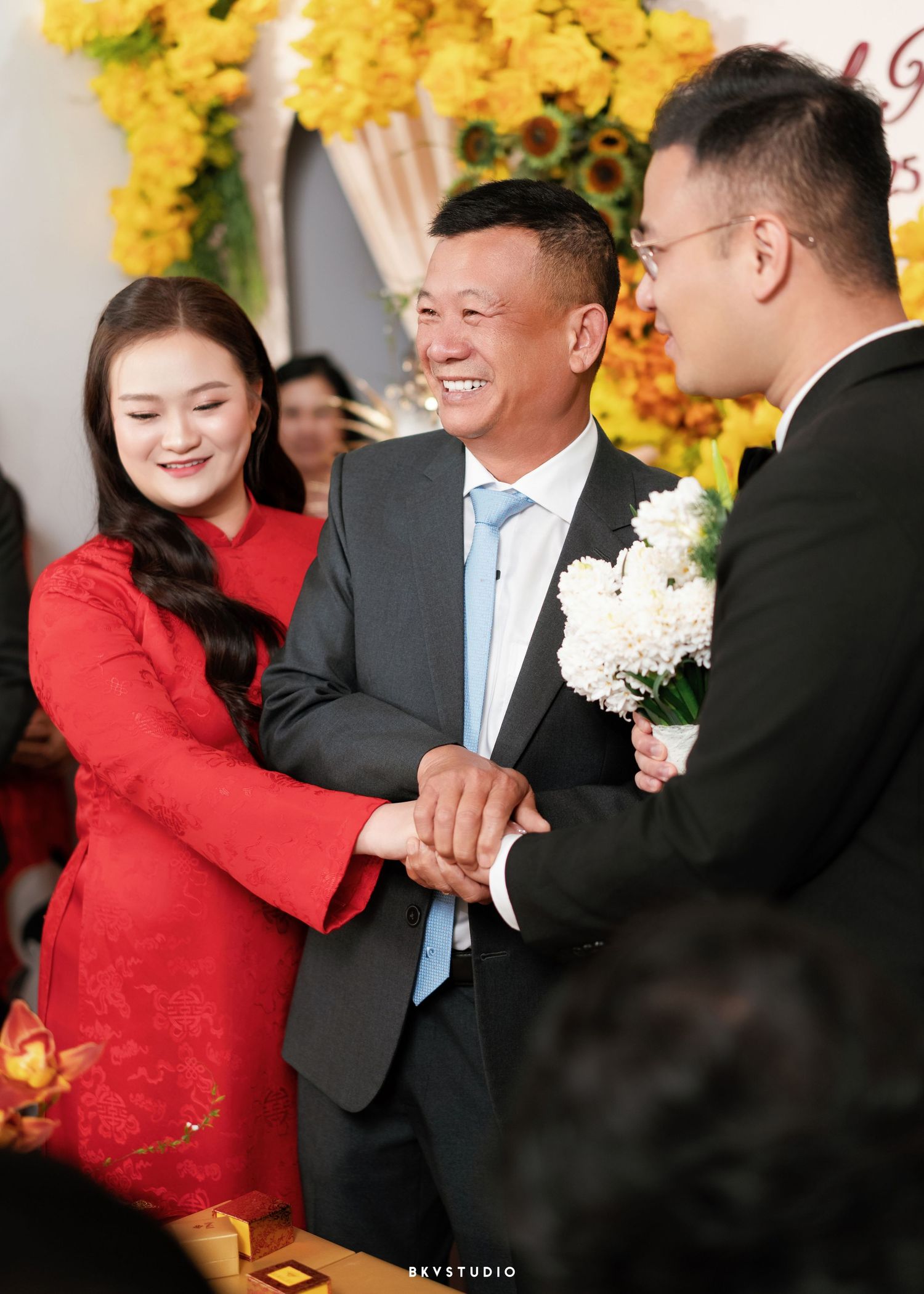 A smiling father joins hands with his daughter in a red ao dai and her groom at a Vietnamese wedding ceremony.