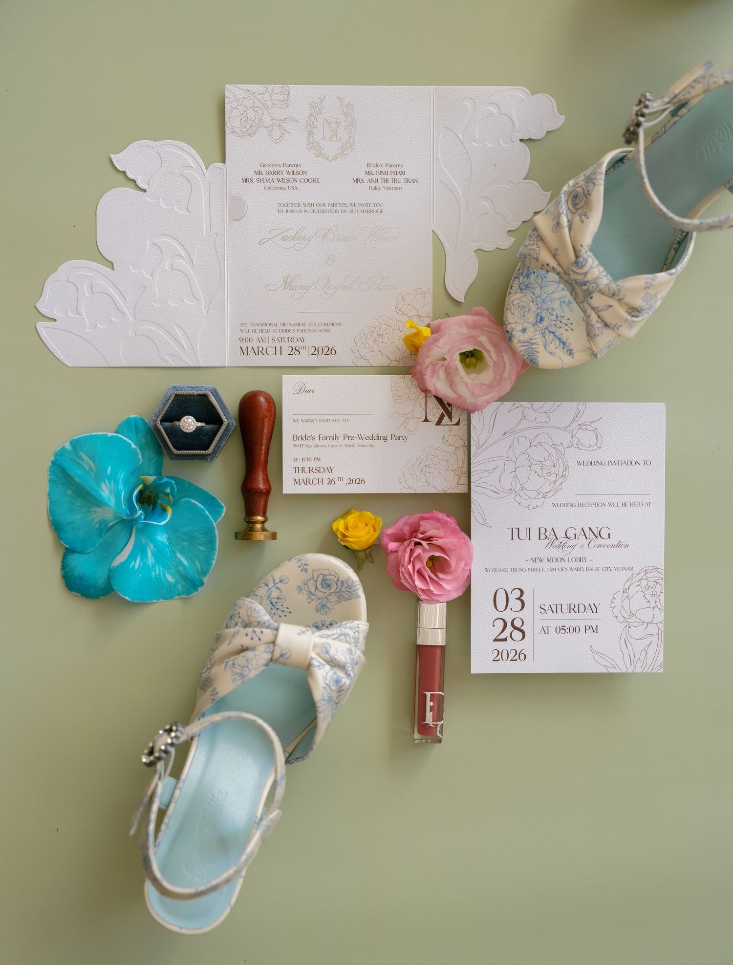 Elegant wedding flat lay with floral invitations, pastel shoes, pink flowers, a ring box, and wax seal on a sage green background.