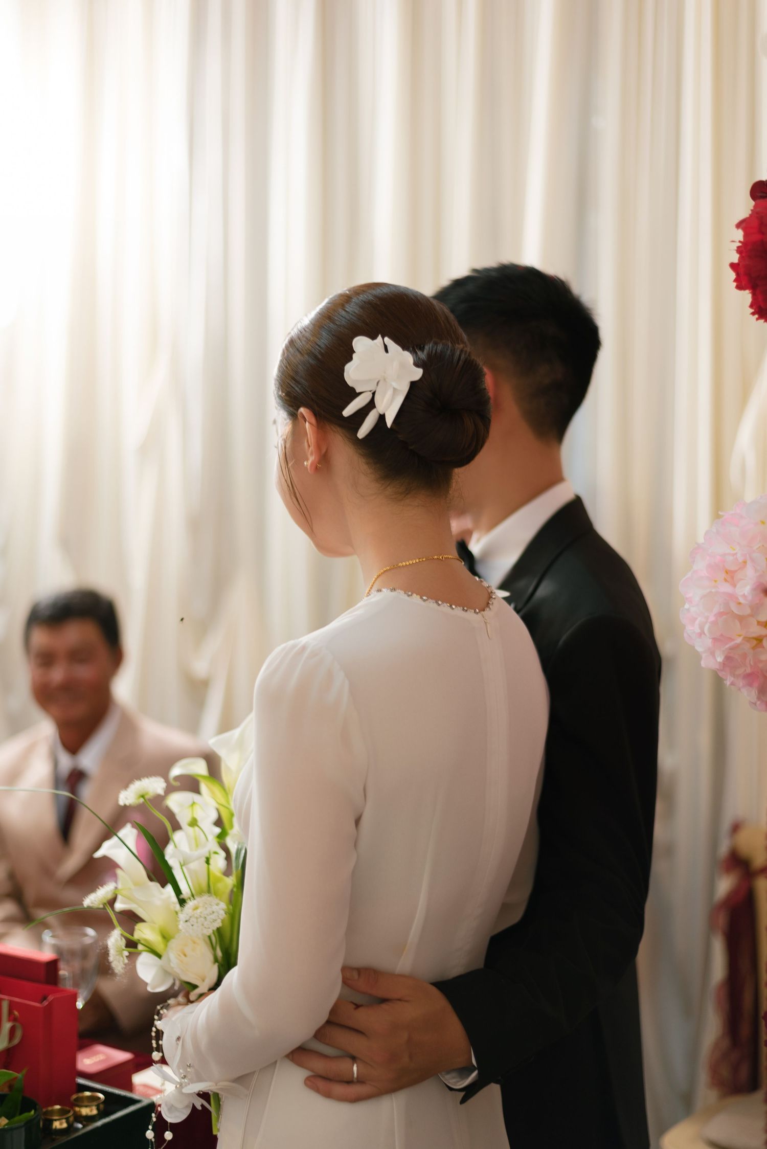 Bride in white ao dai with floral hair accessory stands with groom in black suit at wedding ceremony.