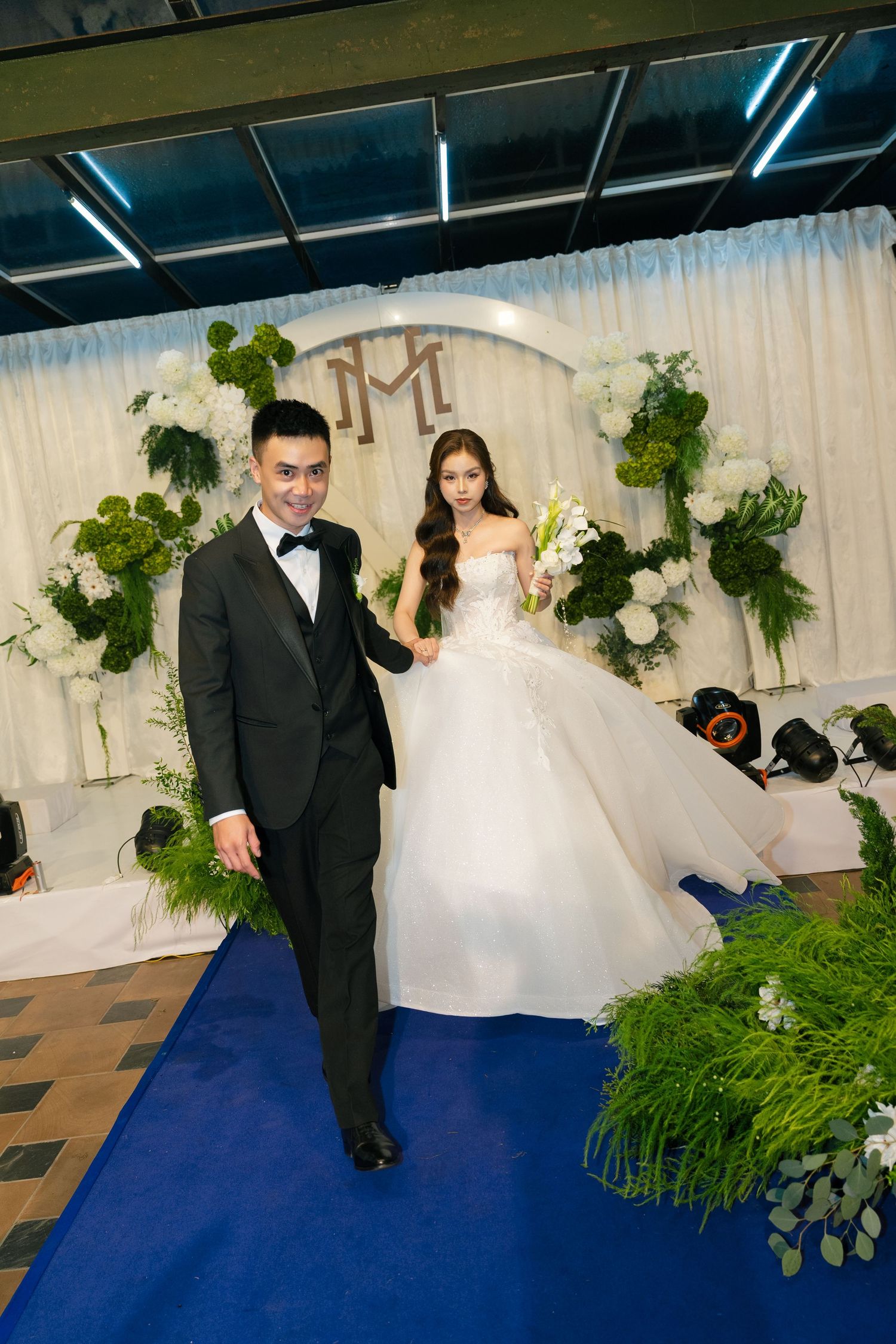 A couple walks down a blue aisle at their wedding, groom in black tuxedo, bride in white ball gown holding white flowers.