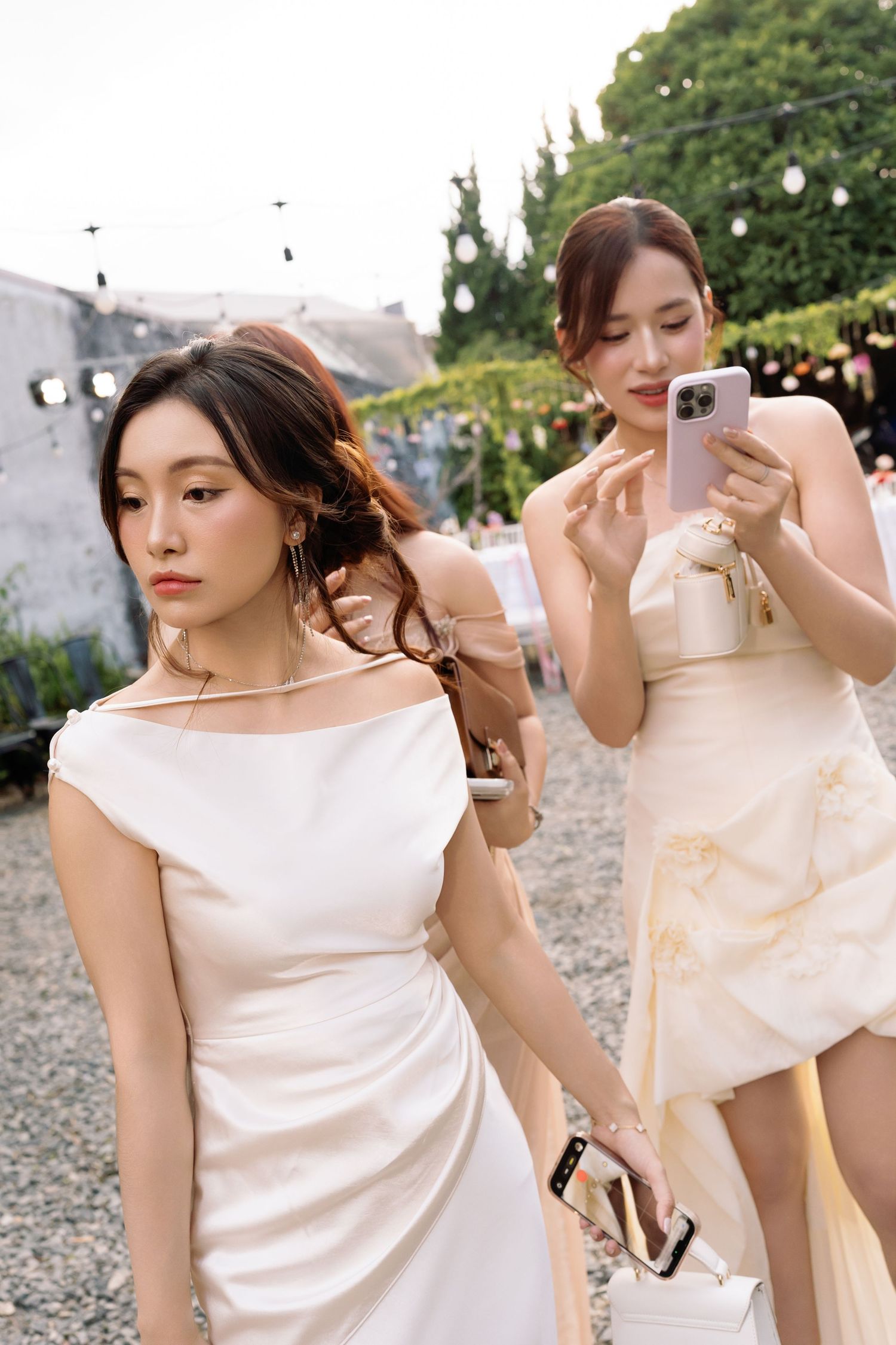 Two elegant women in white dresses at an outdoor event, one taking a photo with a smartphone.