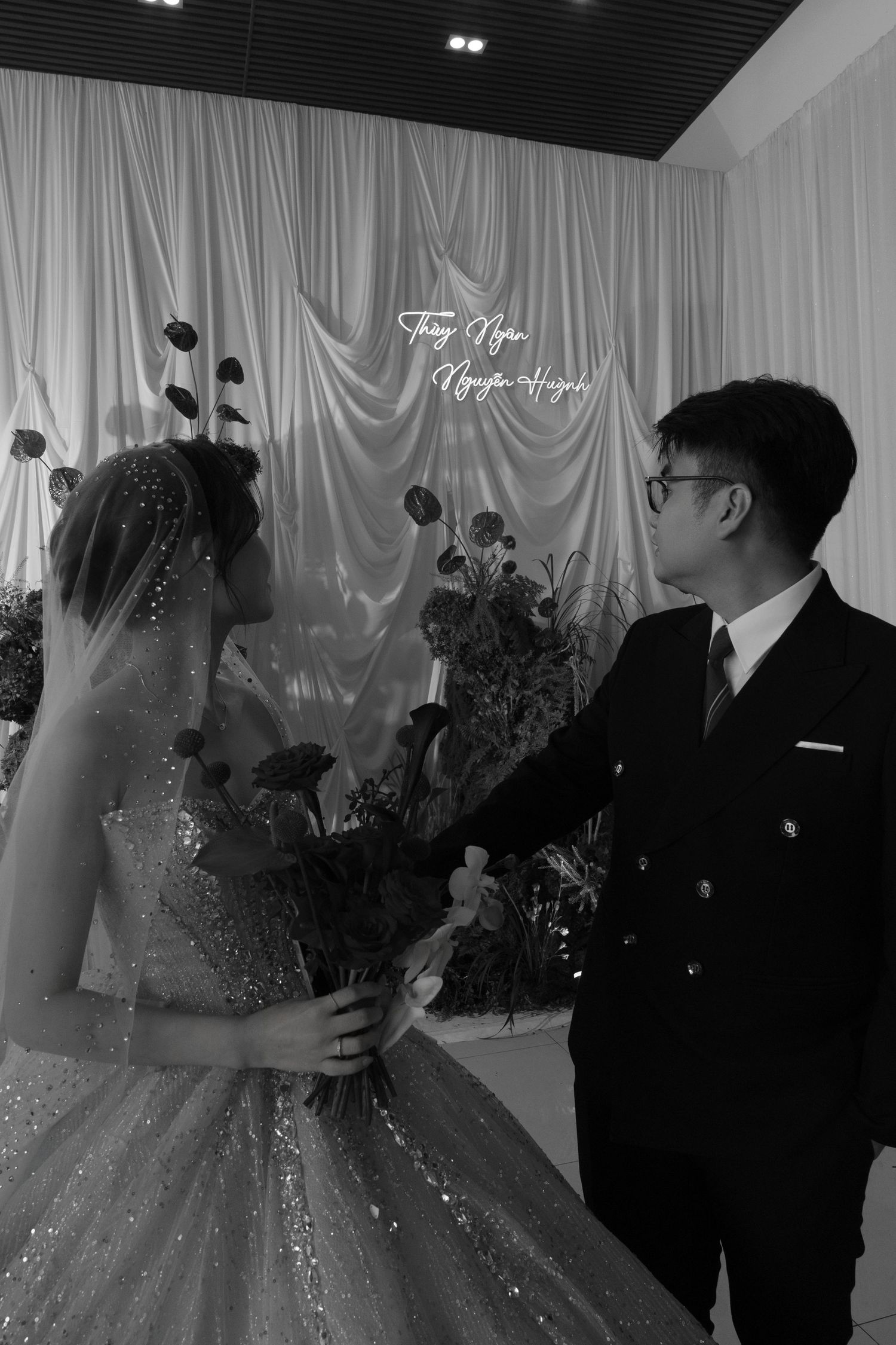 Black and white wedding photo of bride in sparkly gown holding roses, facing groom in suit before draped backdrop.