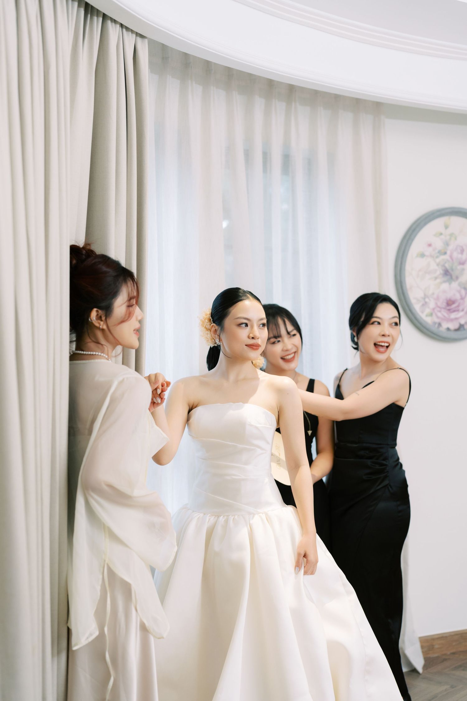 Bride in strapless white gown being helped by bridesmaids in black dresses during wedding preparation.