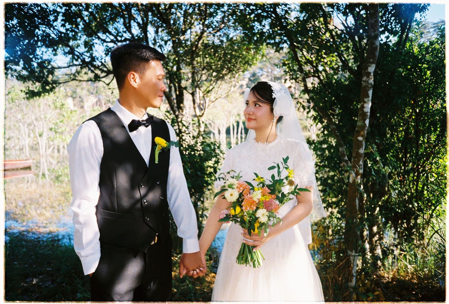 Bride and groom hold hands and smile at each other outdoors, bride holding a colorful bouquet on their wedding day.