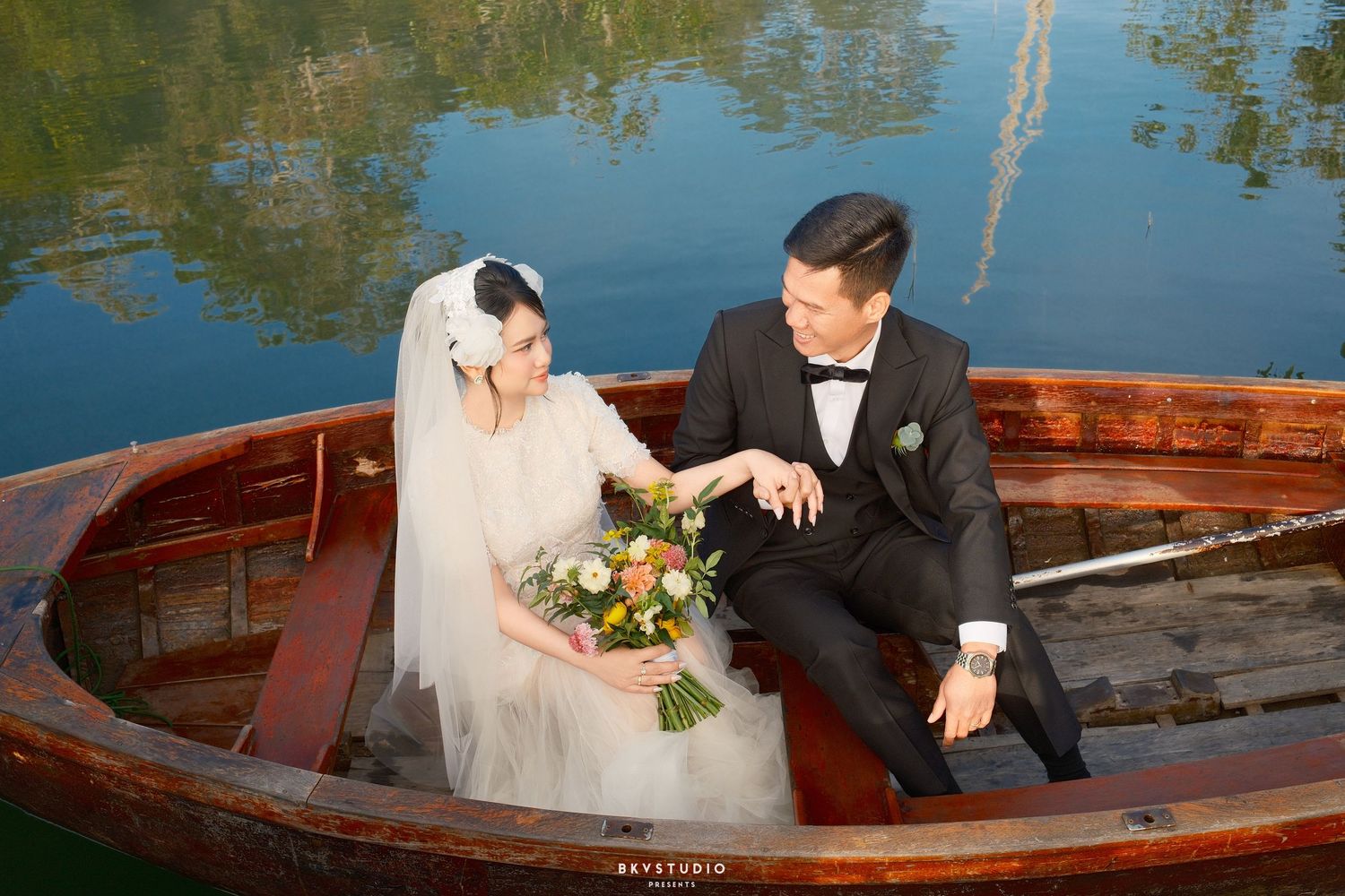 Bride and groom sitting together in a wooden boat, sharing a tender moment with a bridal bouquet on a calm lake.