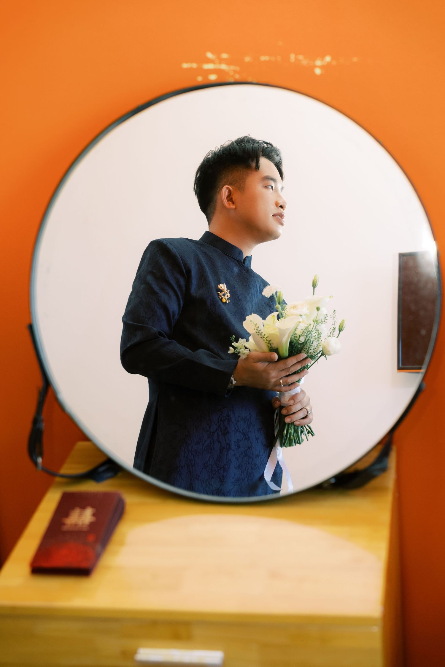 Groom in navy blue traditional attire holds white flower bouquet reflected in round mirror at wedding.