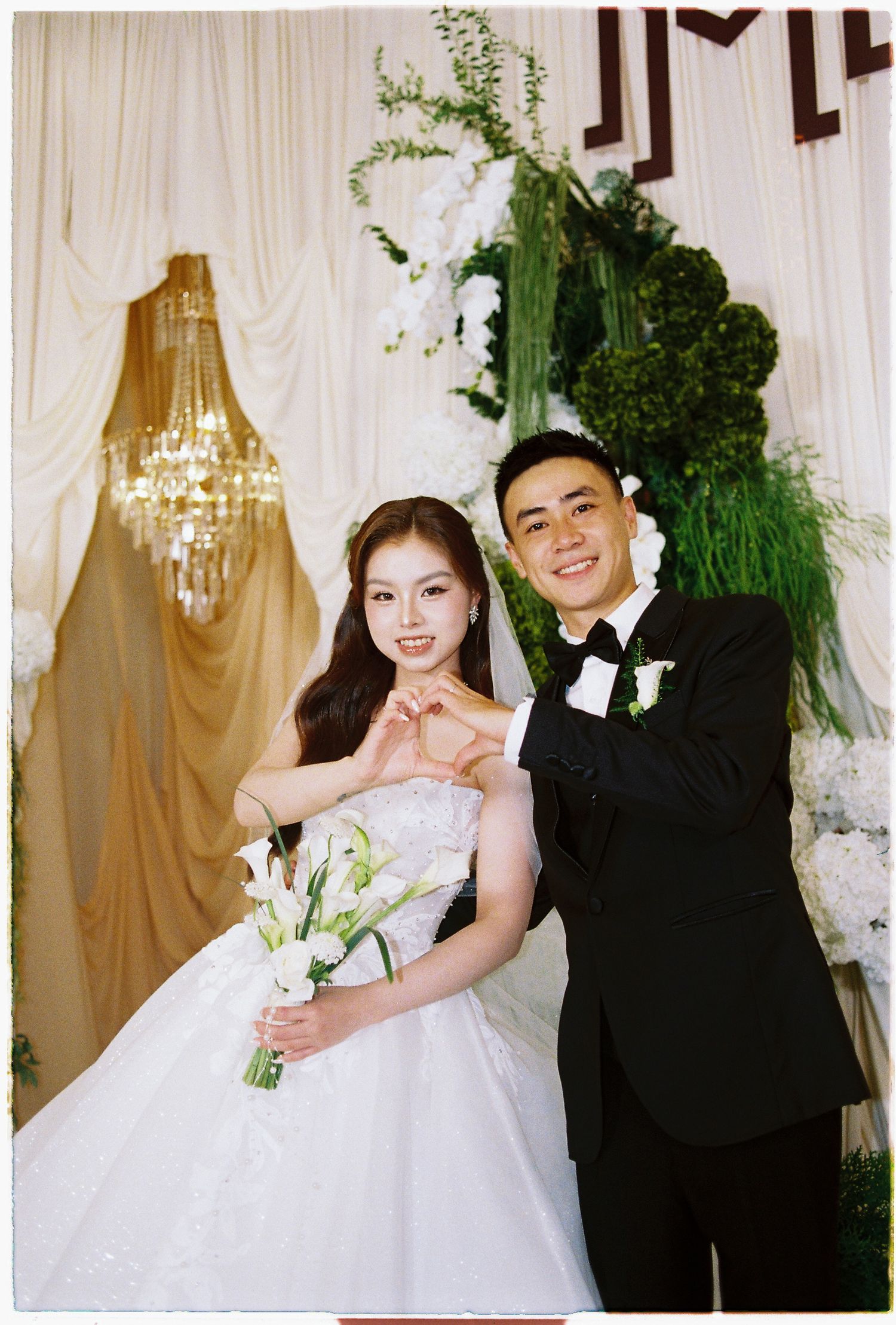 A bride in a white gown and groom in a black tuxedo make a heart shape with their hands at their wedding ceremony.