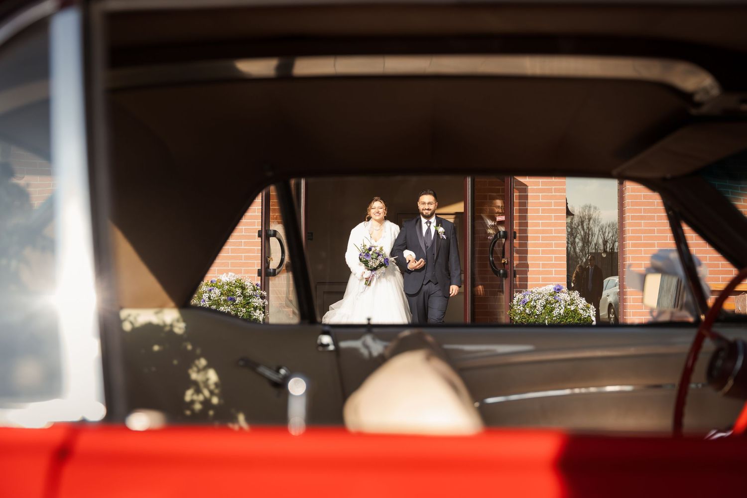 Bride and groom photographed through a vintage red car window on their wedding day.