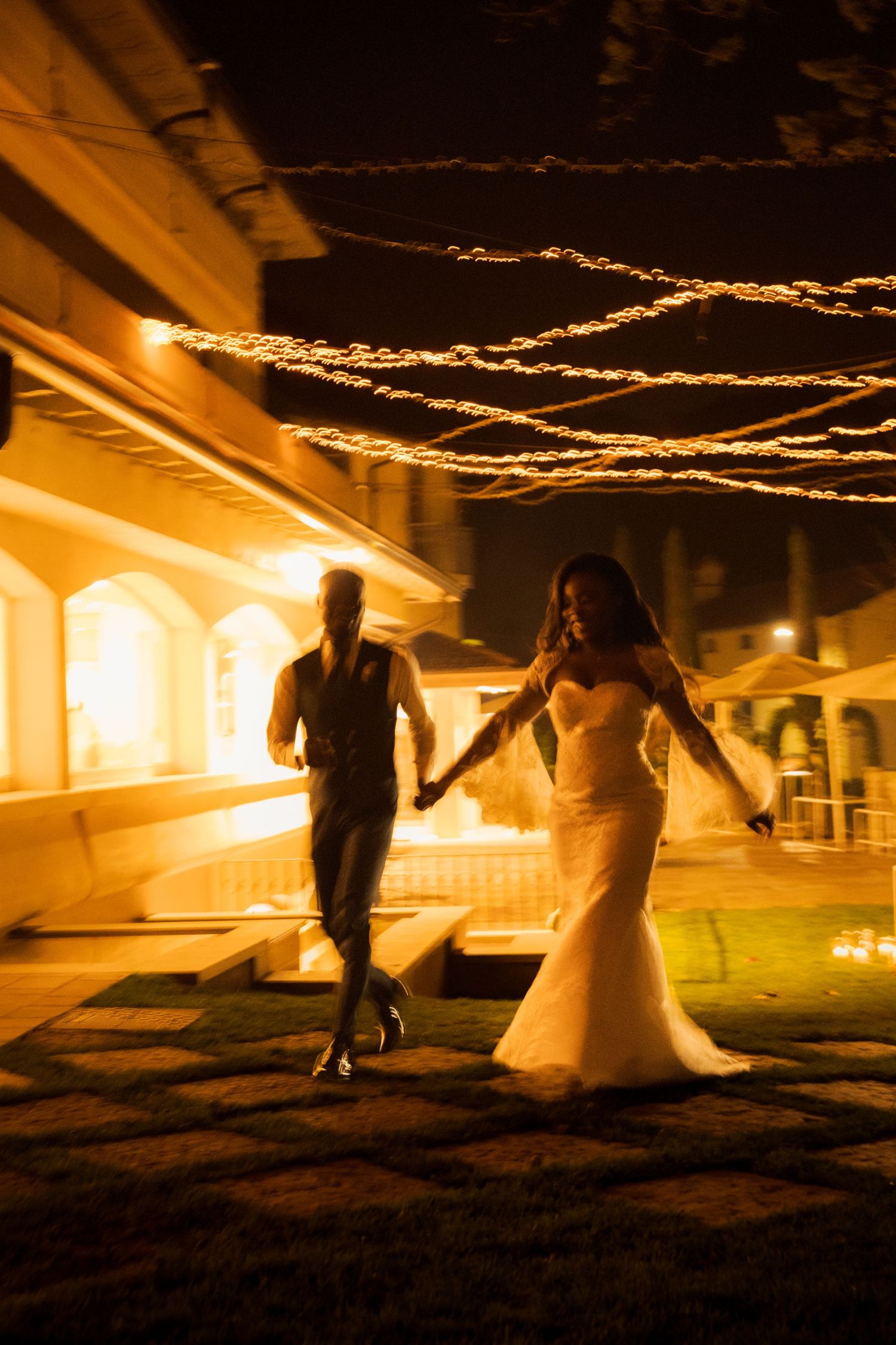 Bride and groom running hand in hand at night under glowing string lights at an outdoor wedding venue.