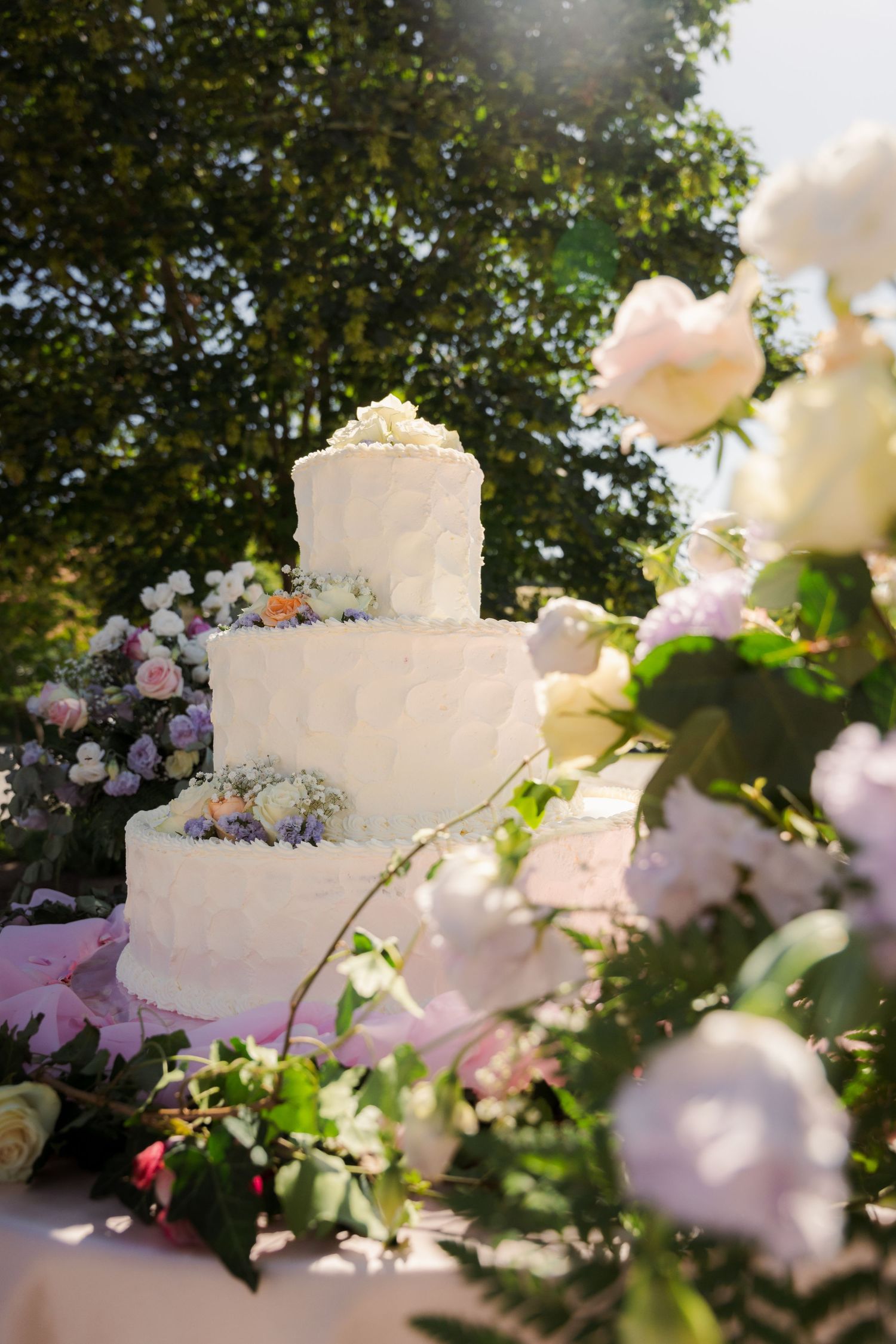 Torta nunziale a tre piano decorata con fiori, allestita all'aperto tra rose bianche e rosa.