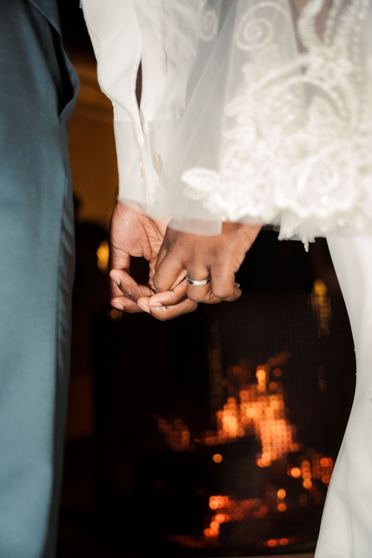 Bride and groom holding hands at wedding ceremony, bride wearing lace dress and wedding ring, warm bokeh lights in background.