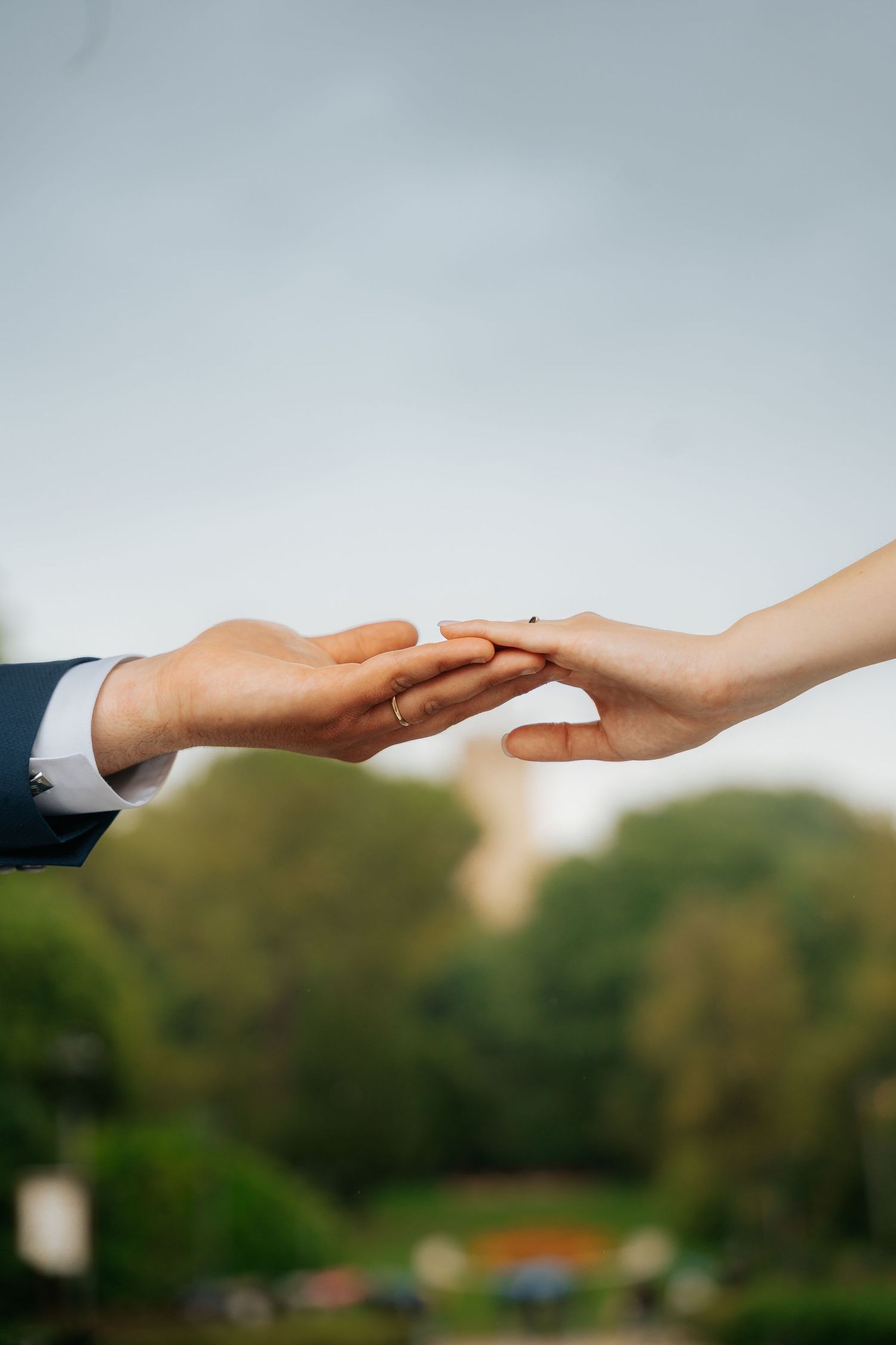 A couple gently holds hands outdoors, wearing wedding rings, with lush green trees and a soft sky in the background.