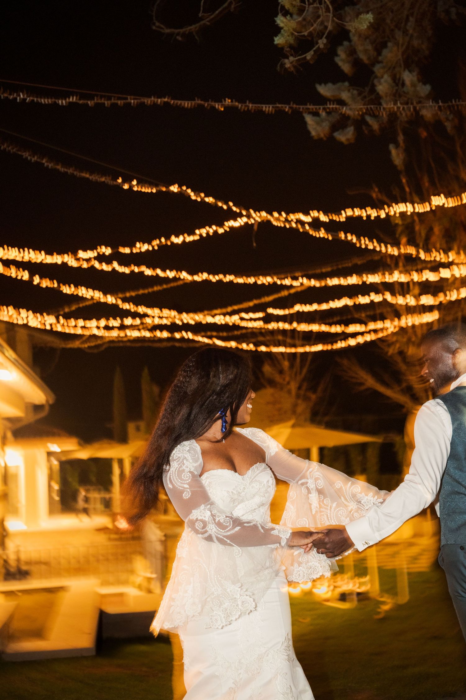 Bride and groom share first dance under glowing string lights at romantic outdoor wedding reception at night.
