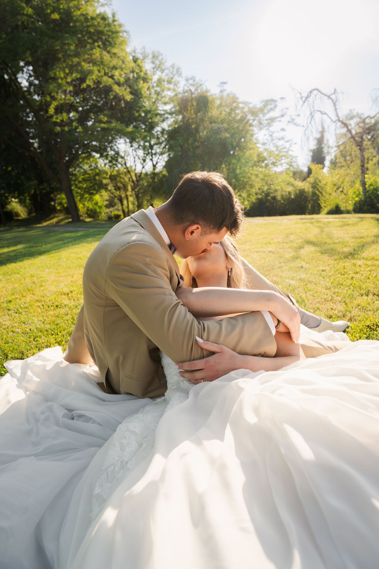 Bride and groom sharing a romantic kiss while sitting on white dress in sunny green park on wedding day.