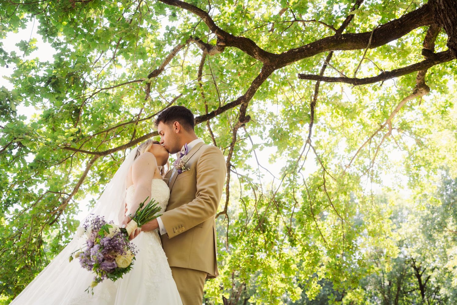 Gli sposi si abbracciano e si baciano sotto i rami di un albero, con il bouquet della sposa in primo piano