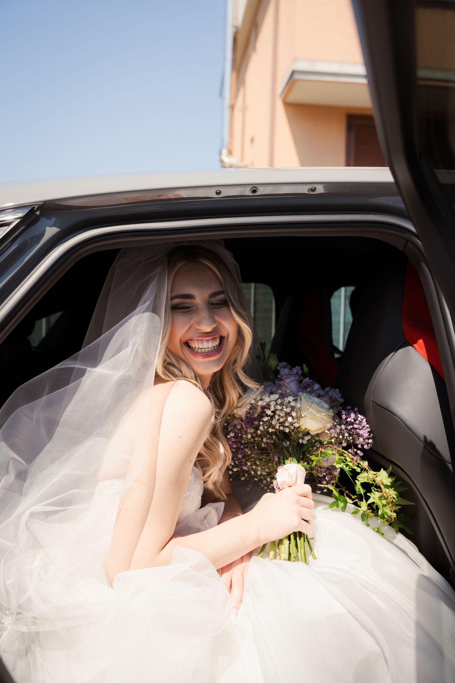 Smiling bride in white gown and veil holds floral bouquet while sitting in a car on her wedding day.