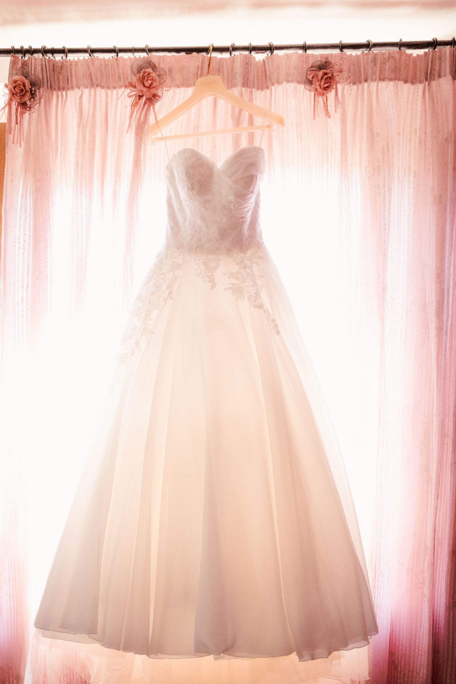 White lace ball gown wedding dress hanging on wooden hanger against pink curtains with rose accents in bridal boutique.