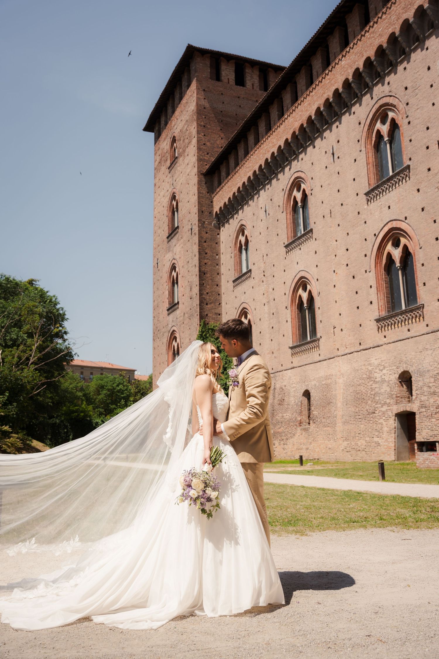 Bride and groom embrace before a historic brick castle on a sunny day, her long veil flowing gracefully in the breeze.