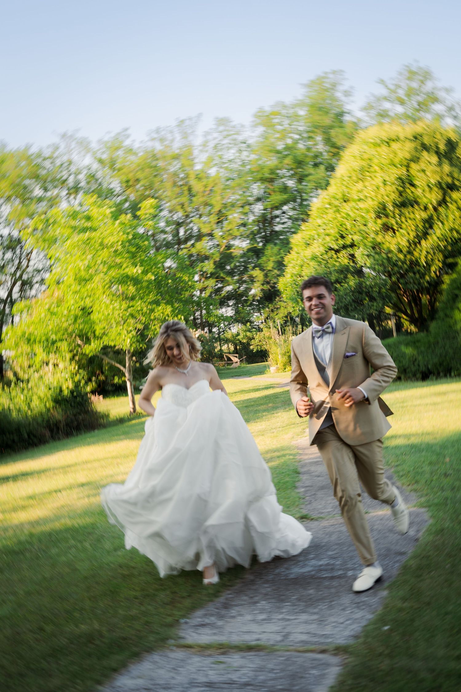 Joyful bride and groom running through lush green garden on their wedding day, bride in white gown, groom in tan suit.