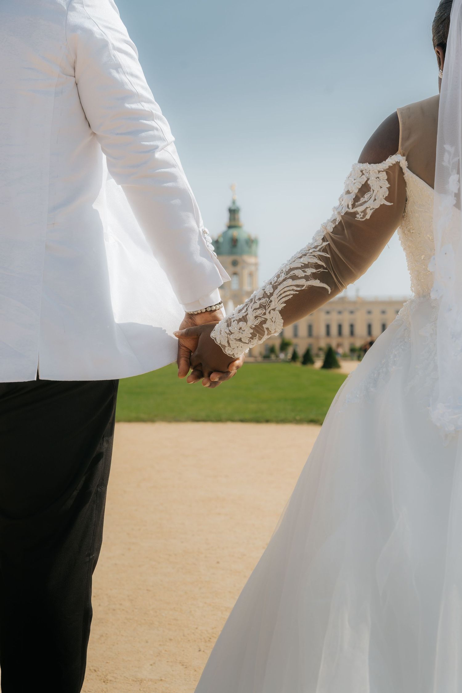 Newlywed couple holding hands in white wedding attire with Charlottenburg Palace in the background.