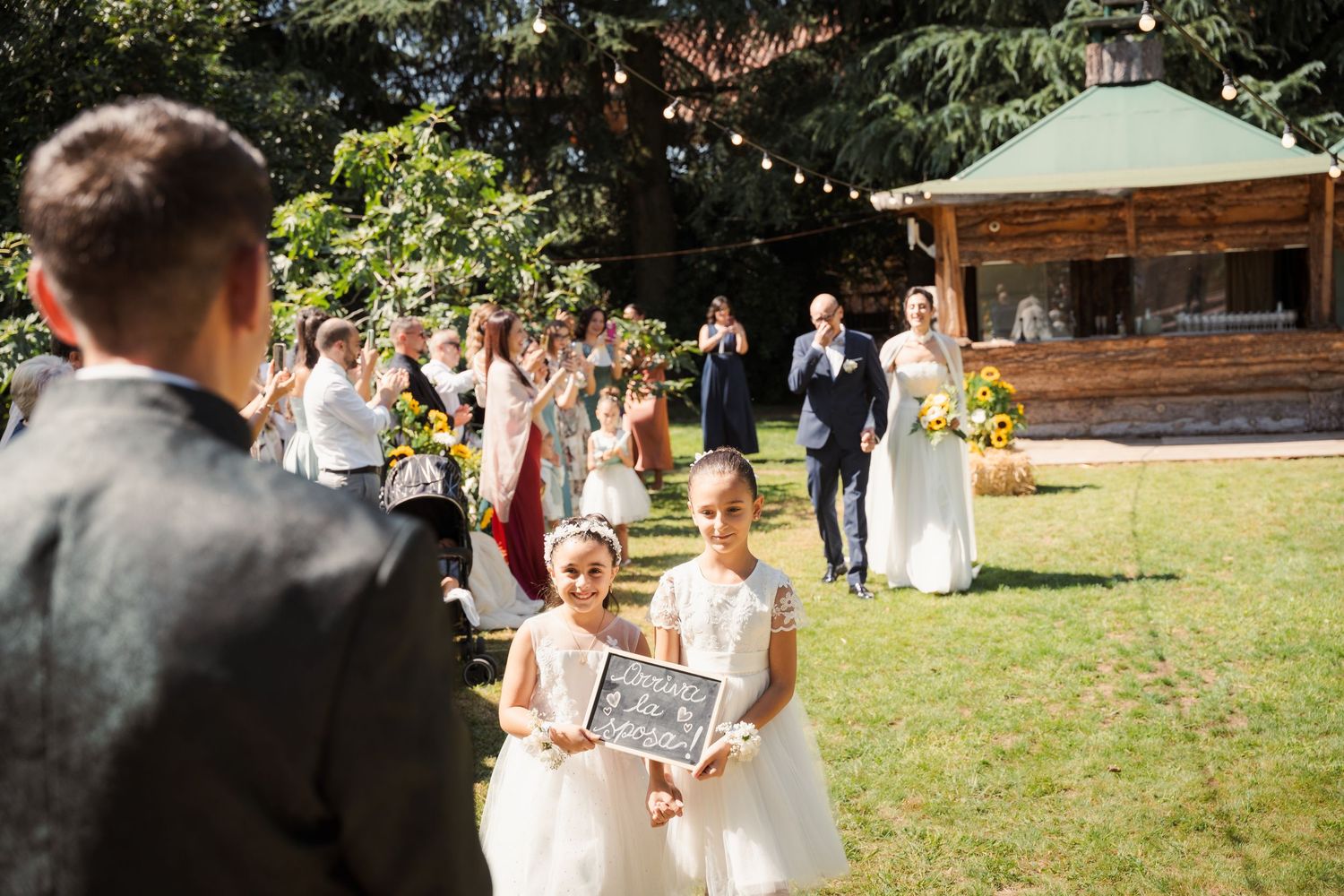Due bambine aprono la cerimonia con un cartello, mentre sullo sfondo la sposa arriva accompagnata dal padre commosso.