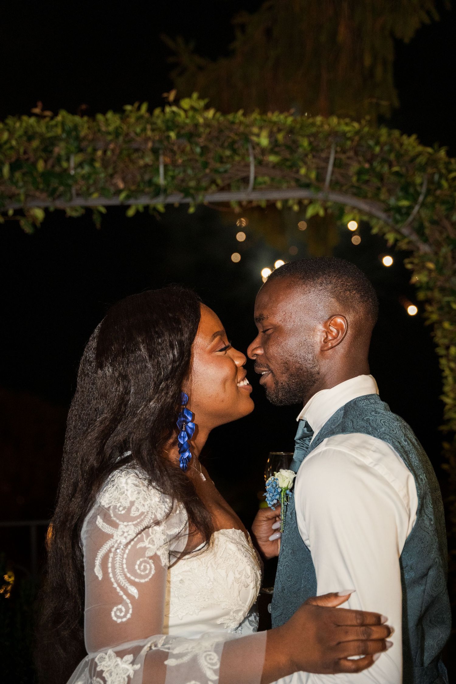 A bride and groom share a romantic moment outdoors at night, surrounded by twinkling string lights.