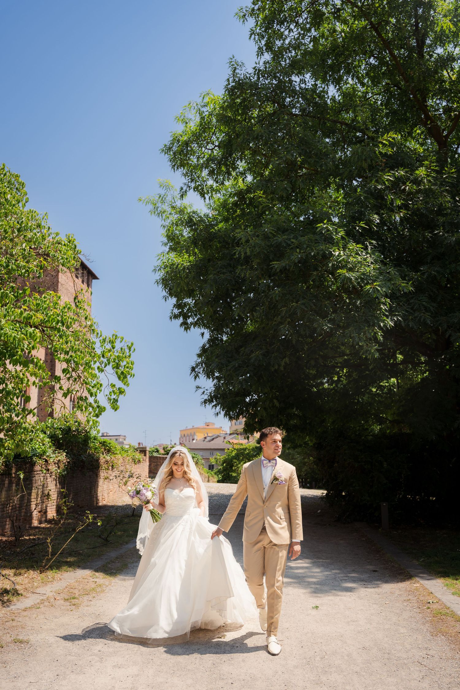 Bride and groom walking hand in hand on a sunny path surrounded by lush green trees on their wedding day.
