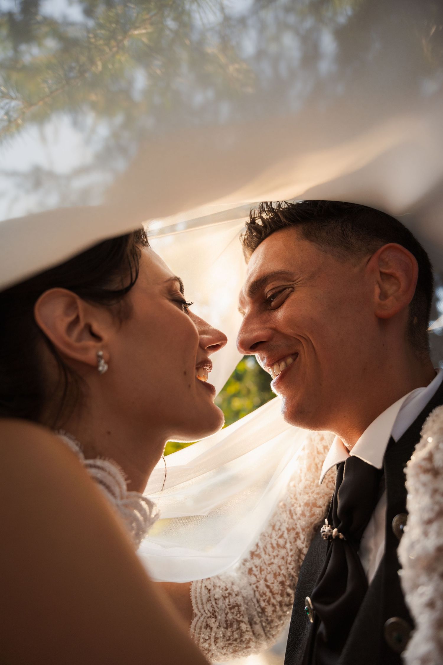 Bride and groom smiling at each other under wedding veil in golden sunlight.