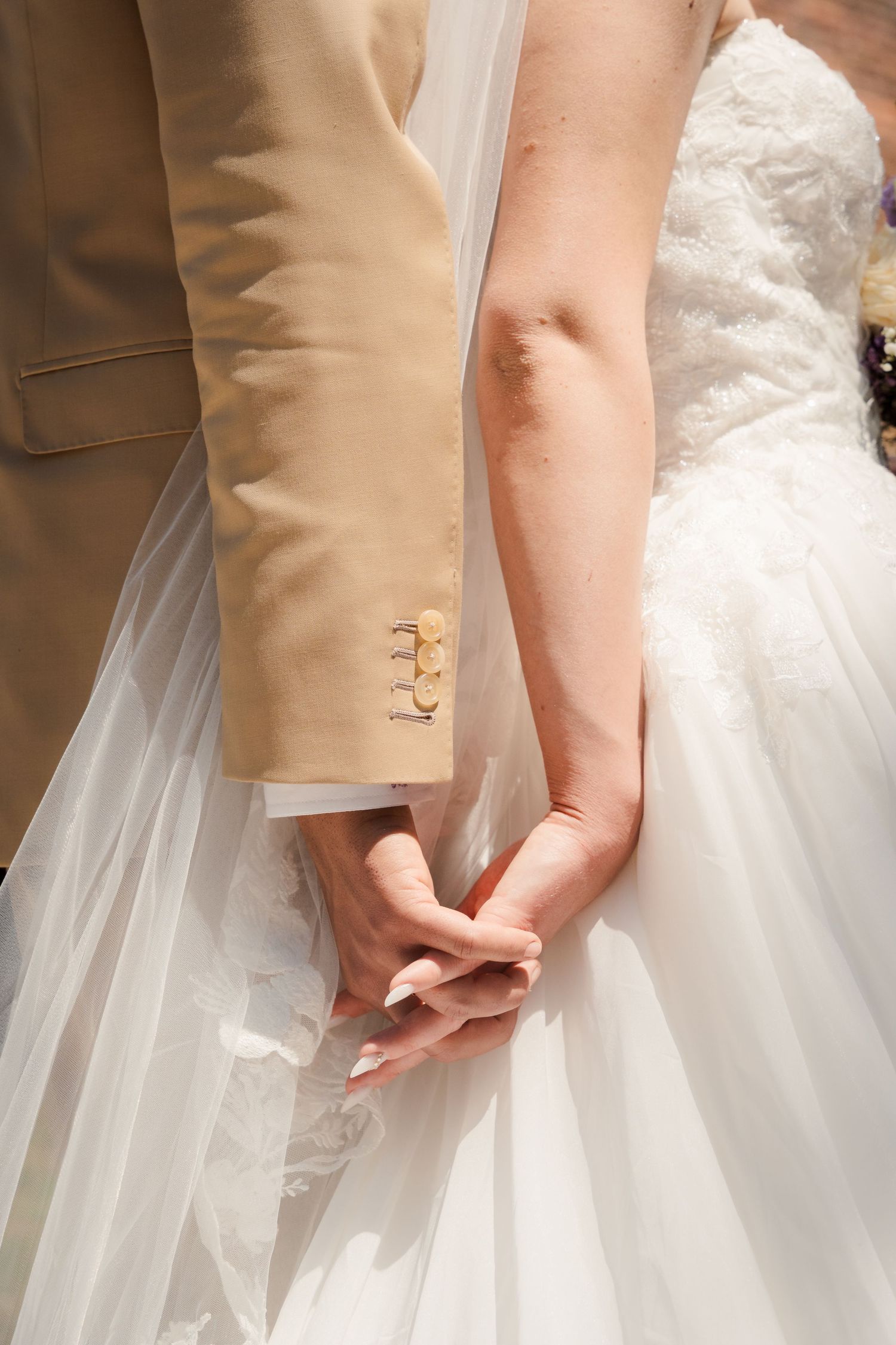 Bride and groom holding hands at wedding, groom in beige suit, bride in white lace gown.