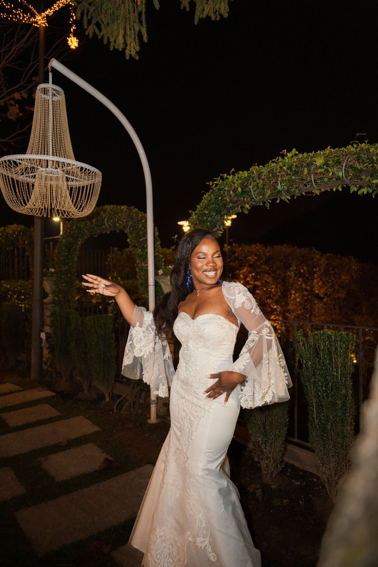 A joyful bride in a white lace mermaid gown with bell sleeves dances outdoors at night near a decorative chandelier.