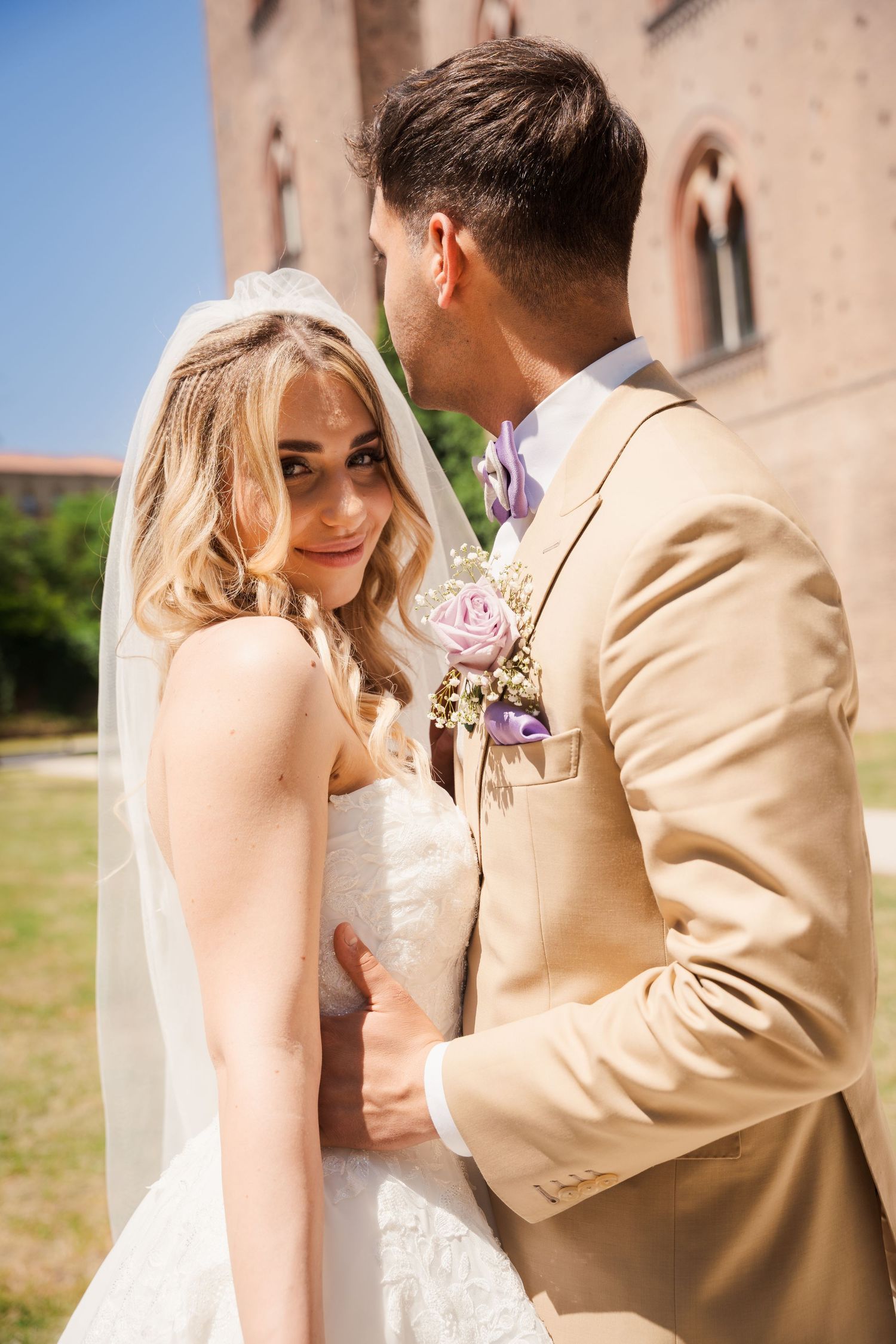 A bride in a white lace dress and veil smiles at the camera while a groom in a beige suit holds her close outdoors.