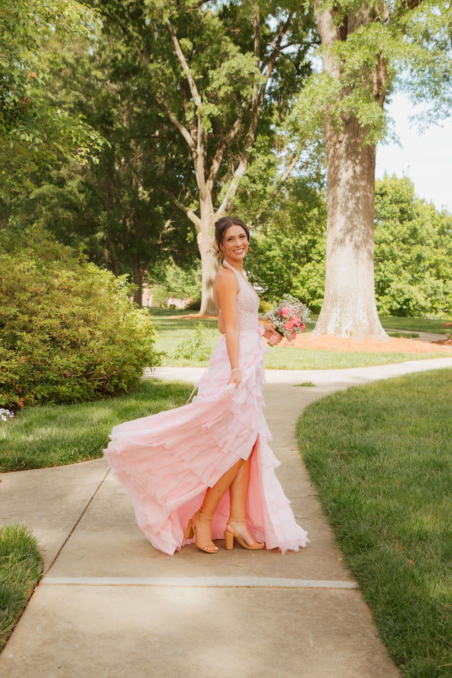 Bride twirling in flowing pink wedding gown on garden pathway, holding bouquet with lush green trees behind her.