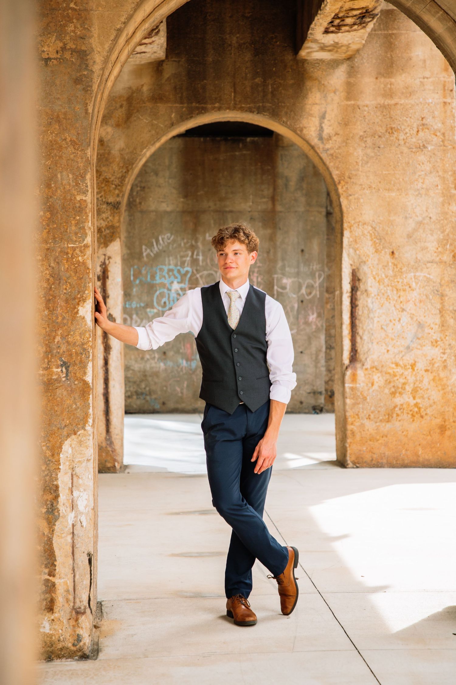 Elegant young man in dark vest poses under stone arches with graffiti in background.
