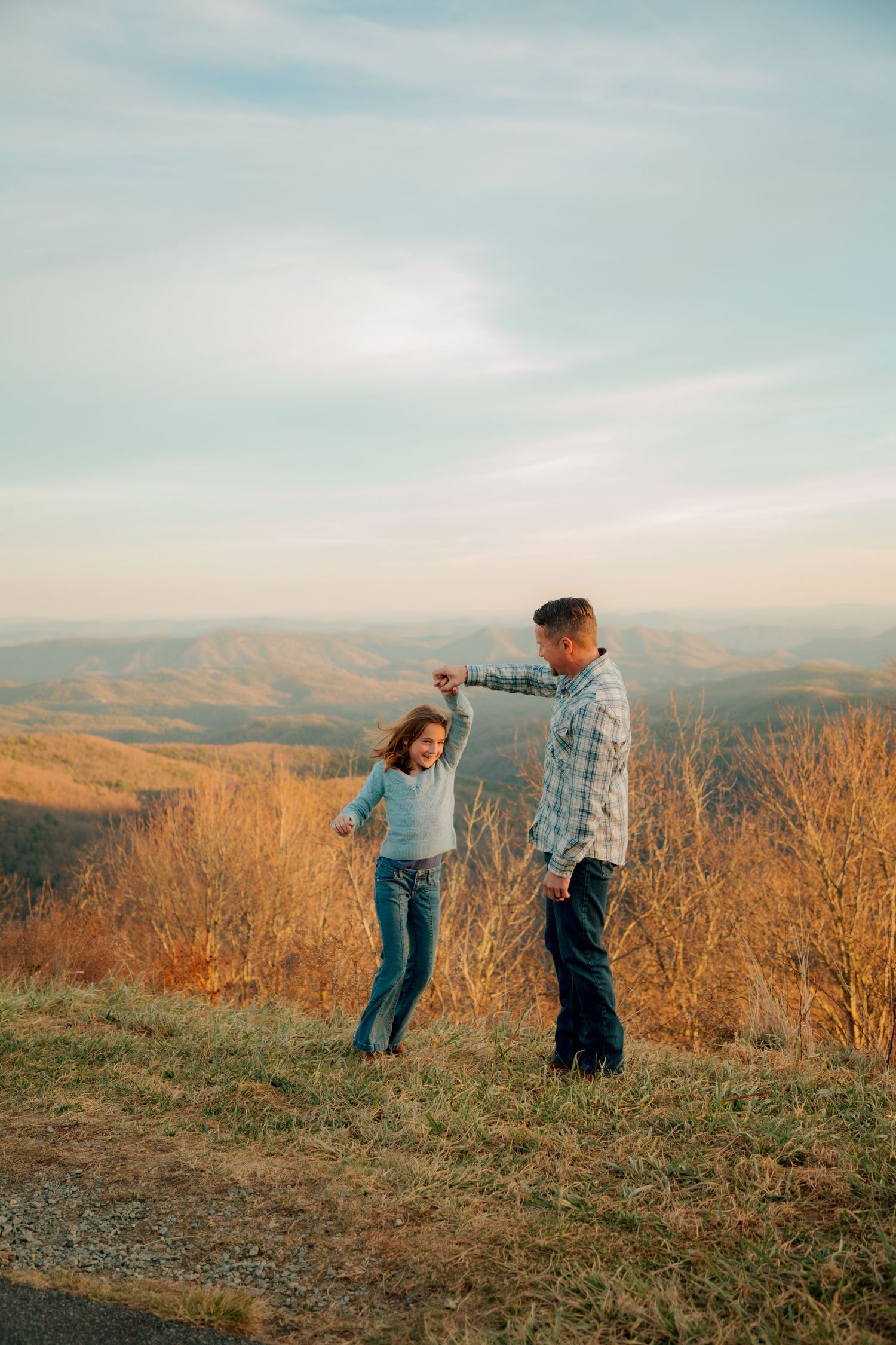 A father in a plaid shirt twirls his young daughter on a hilltop with stunning autumn mountain views behind them.