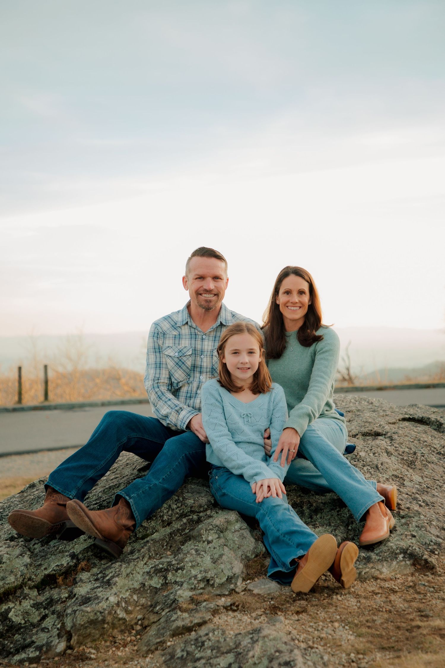 Happy family of three sits on a rock together smiling with a mountain vista behind them.