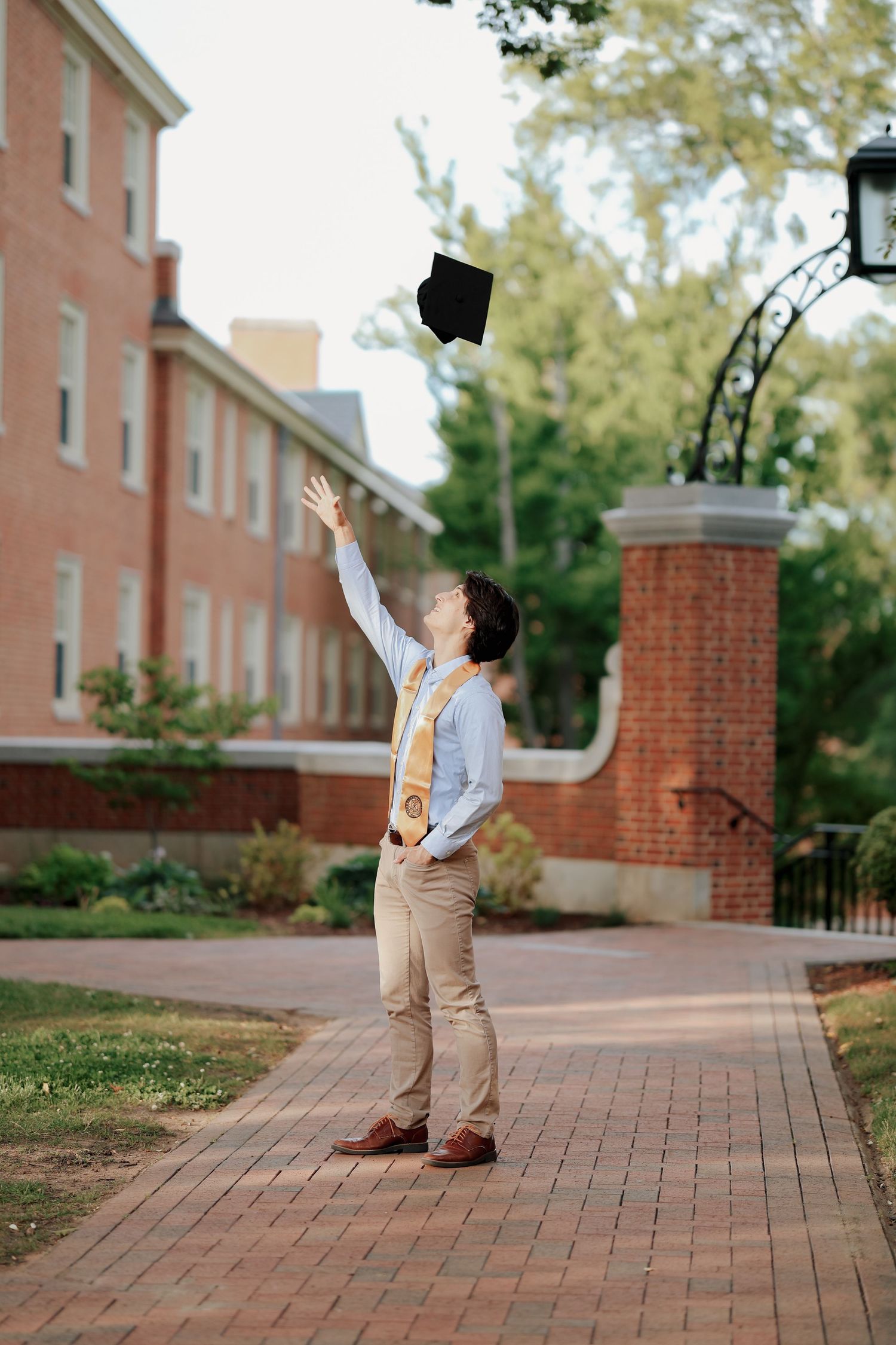 Graduate tosses black mortarboard cap in air on brick university pathway near iron gate.