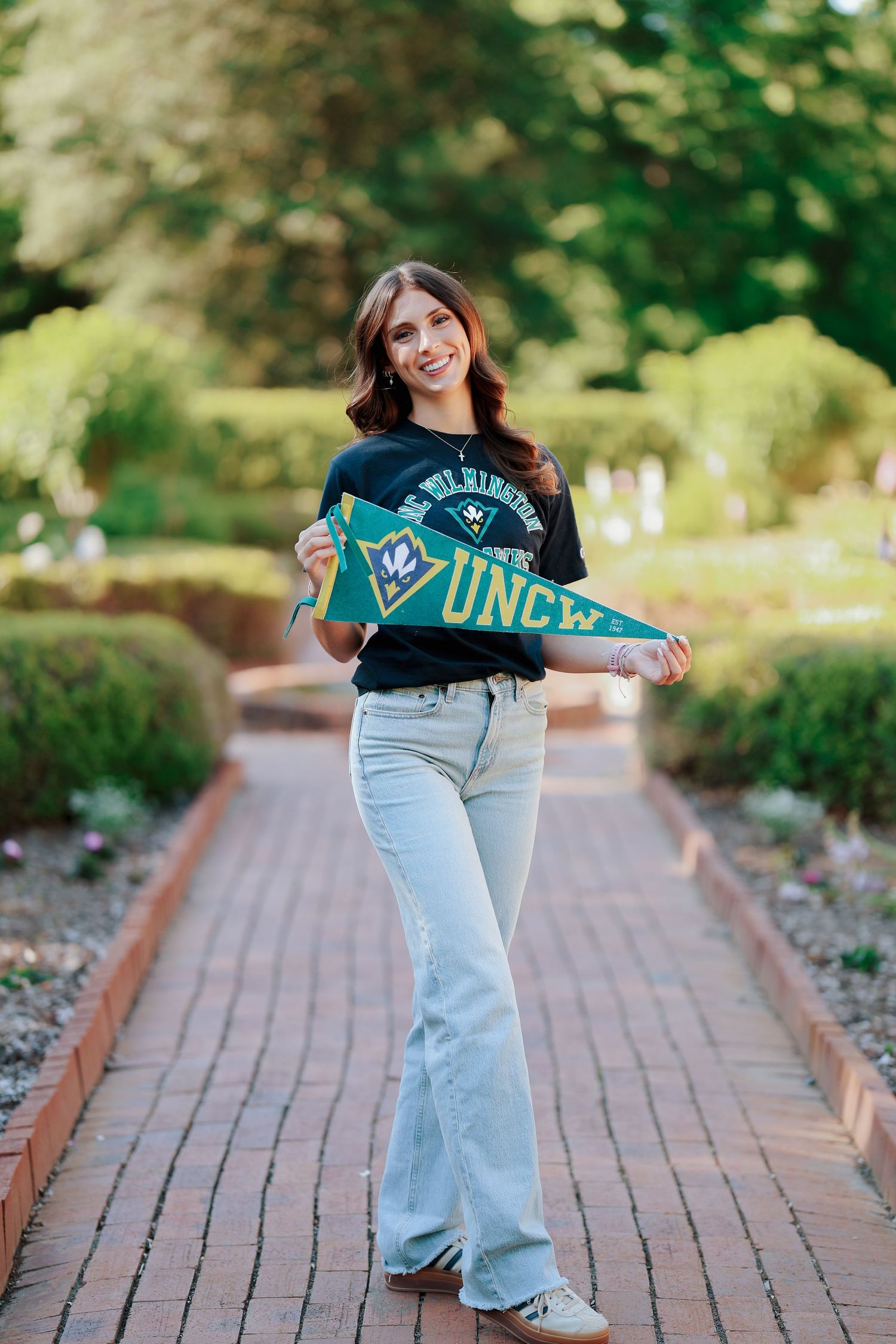 Young woman holding a UNCW pennant flag while smiling on a brick path in a green garden setting.