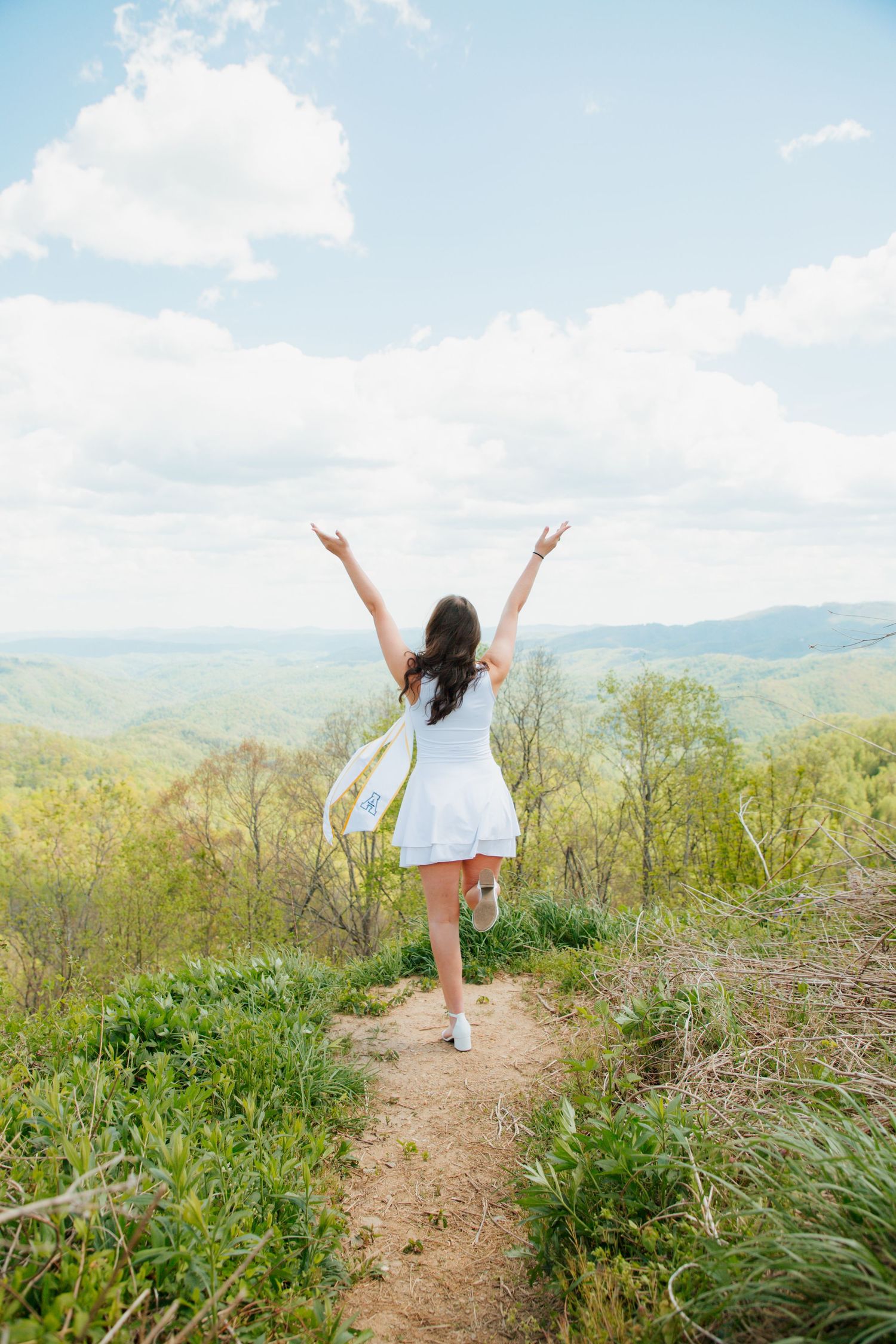 Woman in white dress with arms raised celebrating on mountain trail with green hills and blue sky.