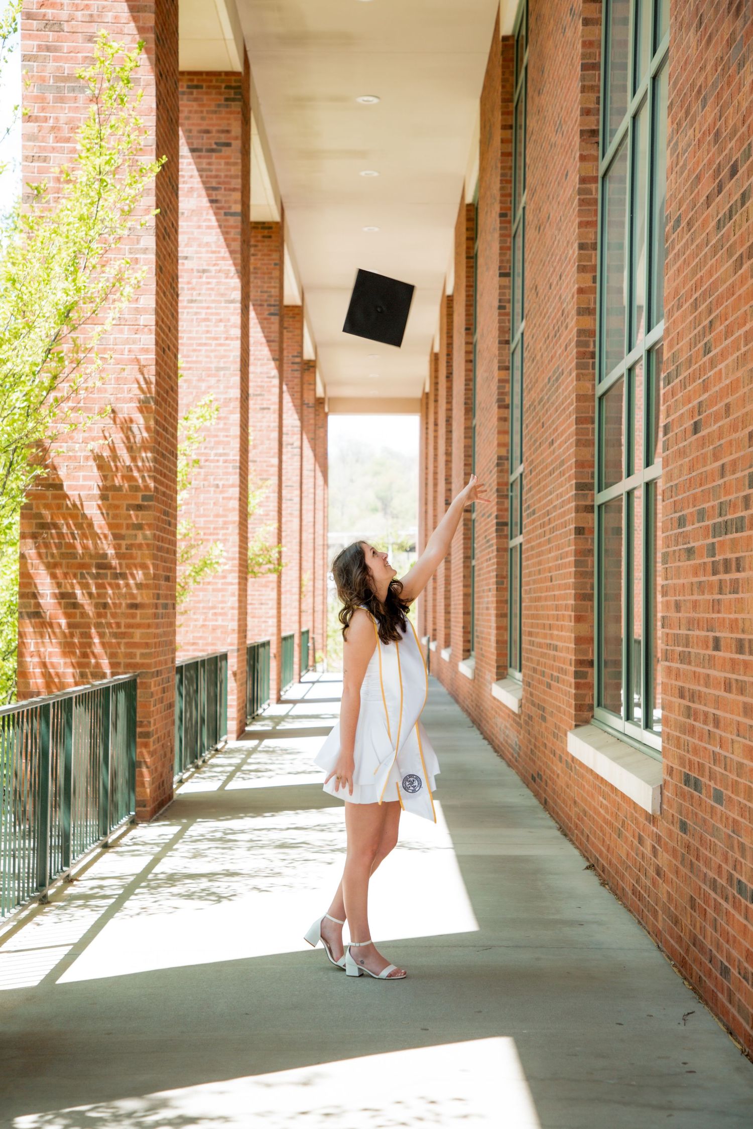 Female graduate in white dress tossing black cap in brick building hallway at Appalachian State University.