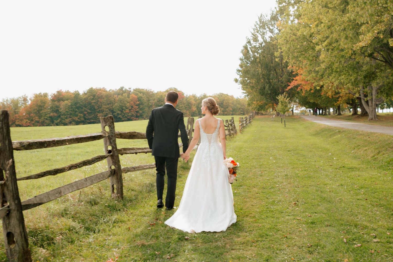Bride and groom walking hand in hand along a rustic fence in autumn.