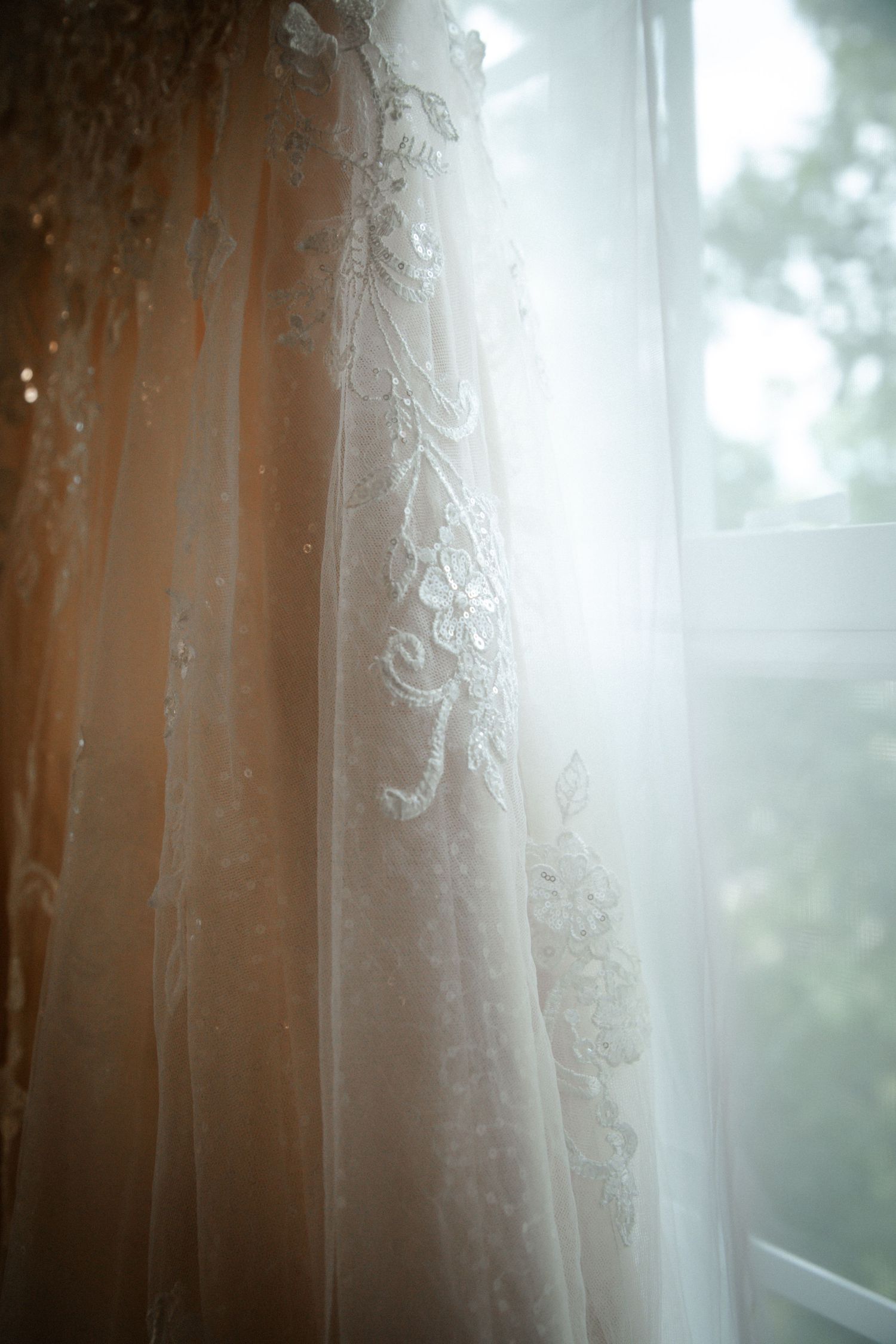 Close-up detail of intricate lace embroidery on blush wedding gown by window.