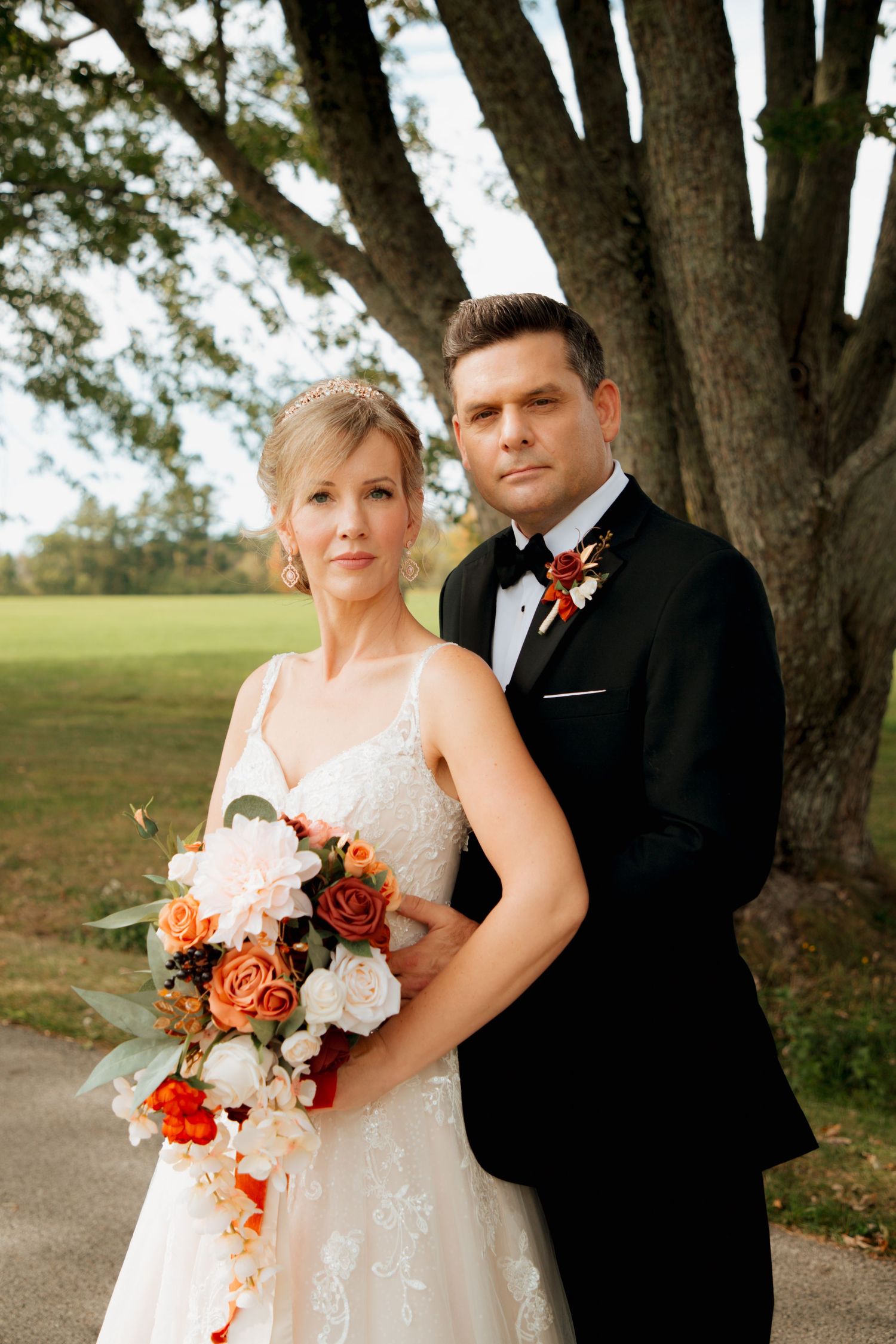 Bride and groom pose formally under a large tree, bride holding a cascading autumn floral bouquet.